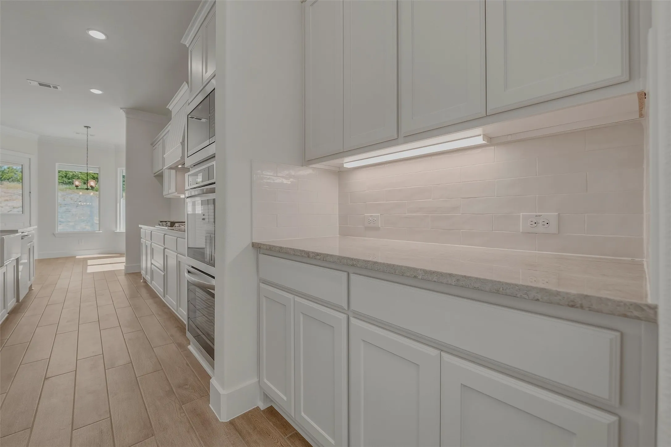Modern kitchen featuring white shaker cabinetry, light-toned countertop, subway tile backsplash with under-cabinet lighting, and integrated stainless steel wall ovens