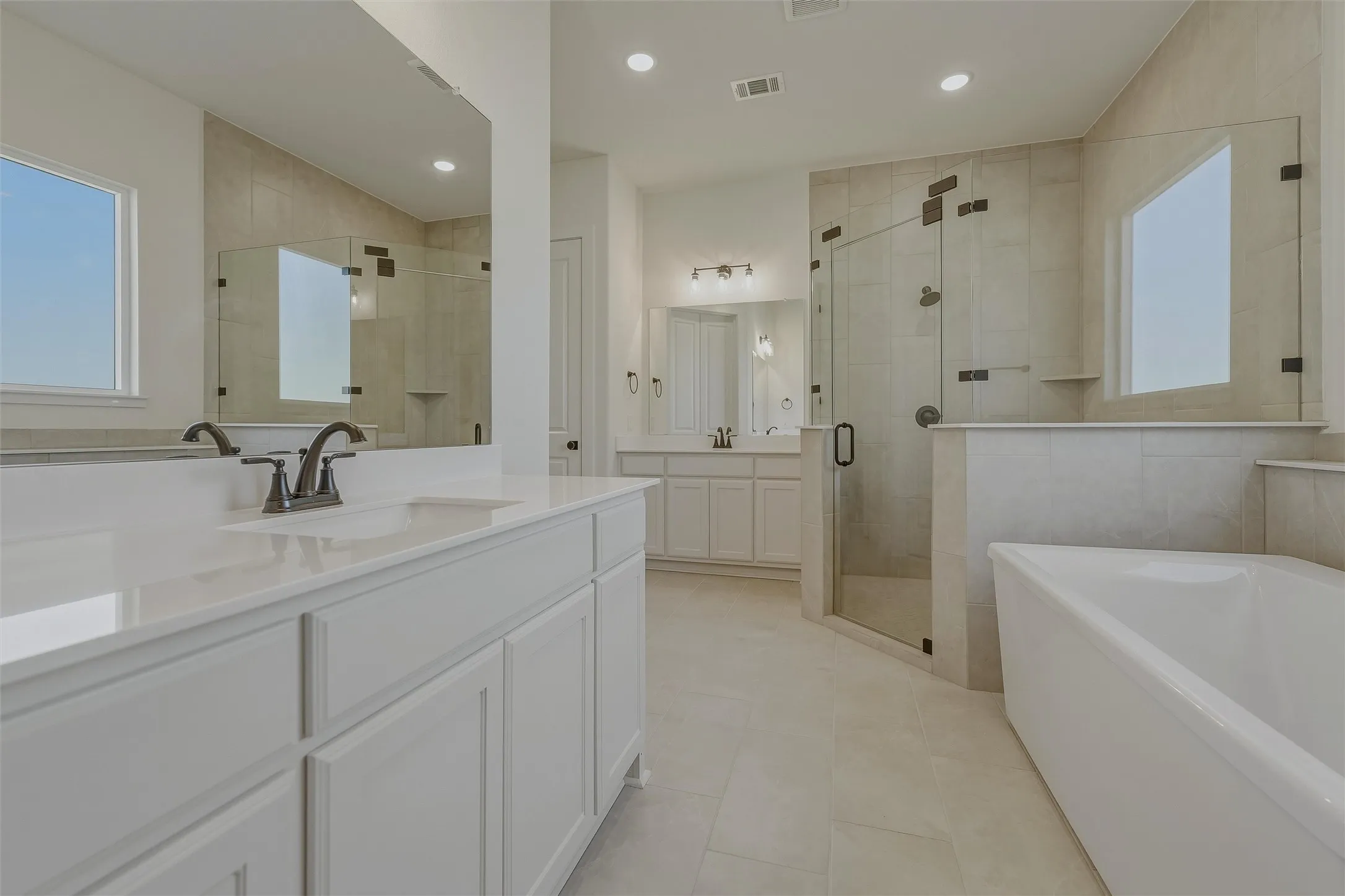 Spacious bathroom featuring a freestanding tub, glass-enclosed shower, dual vanities with white cabinetry, and large format tiled flooring