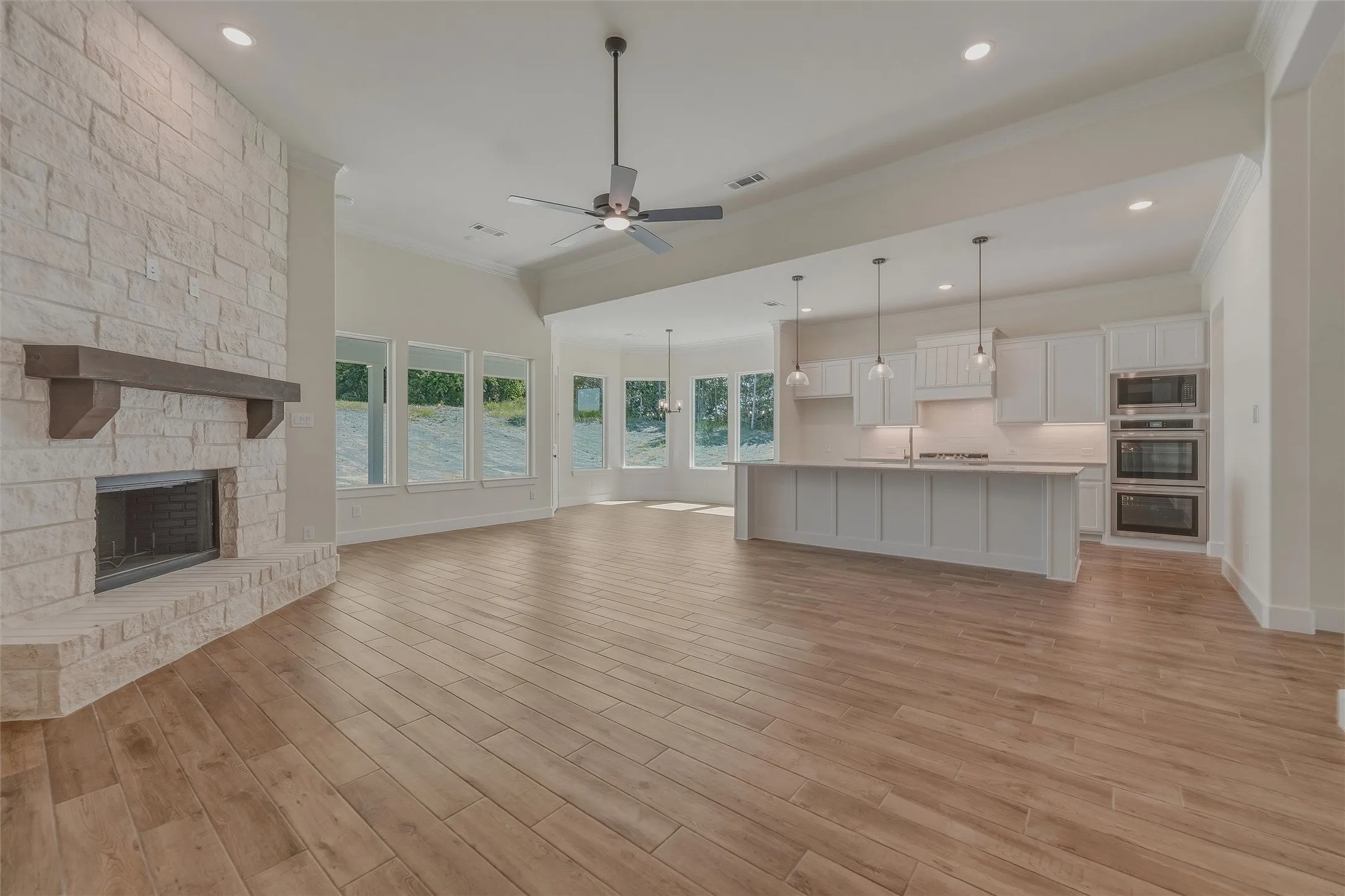 Spacious interior featuring wood-finish flooring, a stone-clad fireplace with a wood mantle, an expansive kitchen island, white cabinetry, and stainless steel built-in appliances
