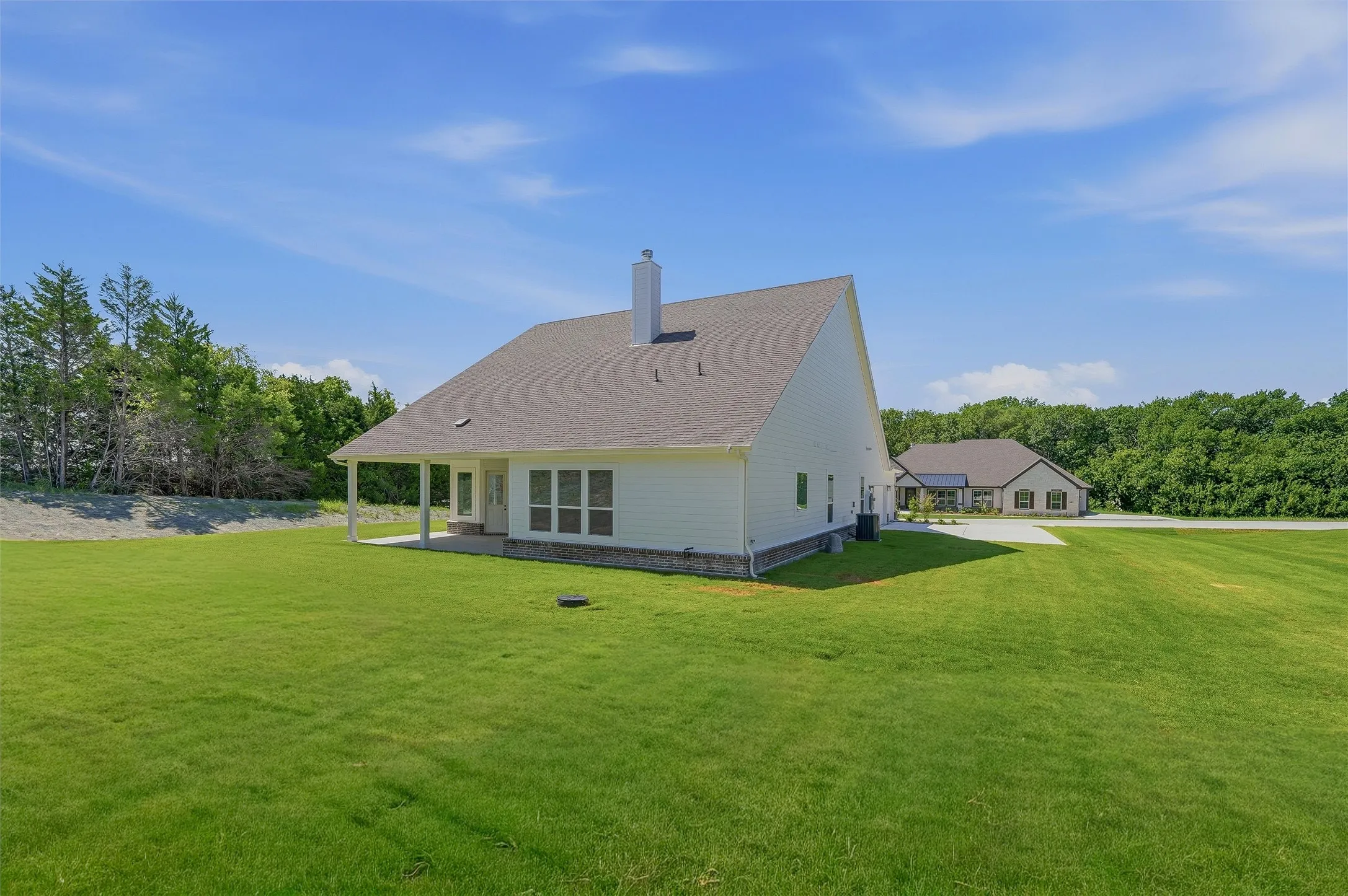Expansive green lawn surrounds a white siding residence with a covered patio, multiple windows, and a prominent chimney