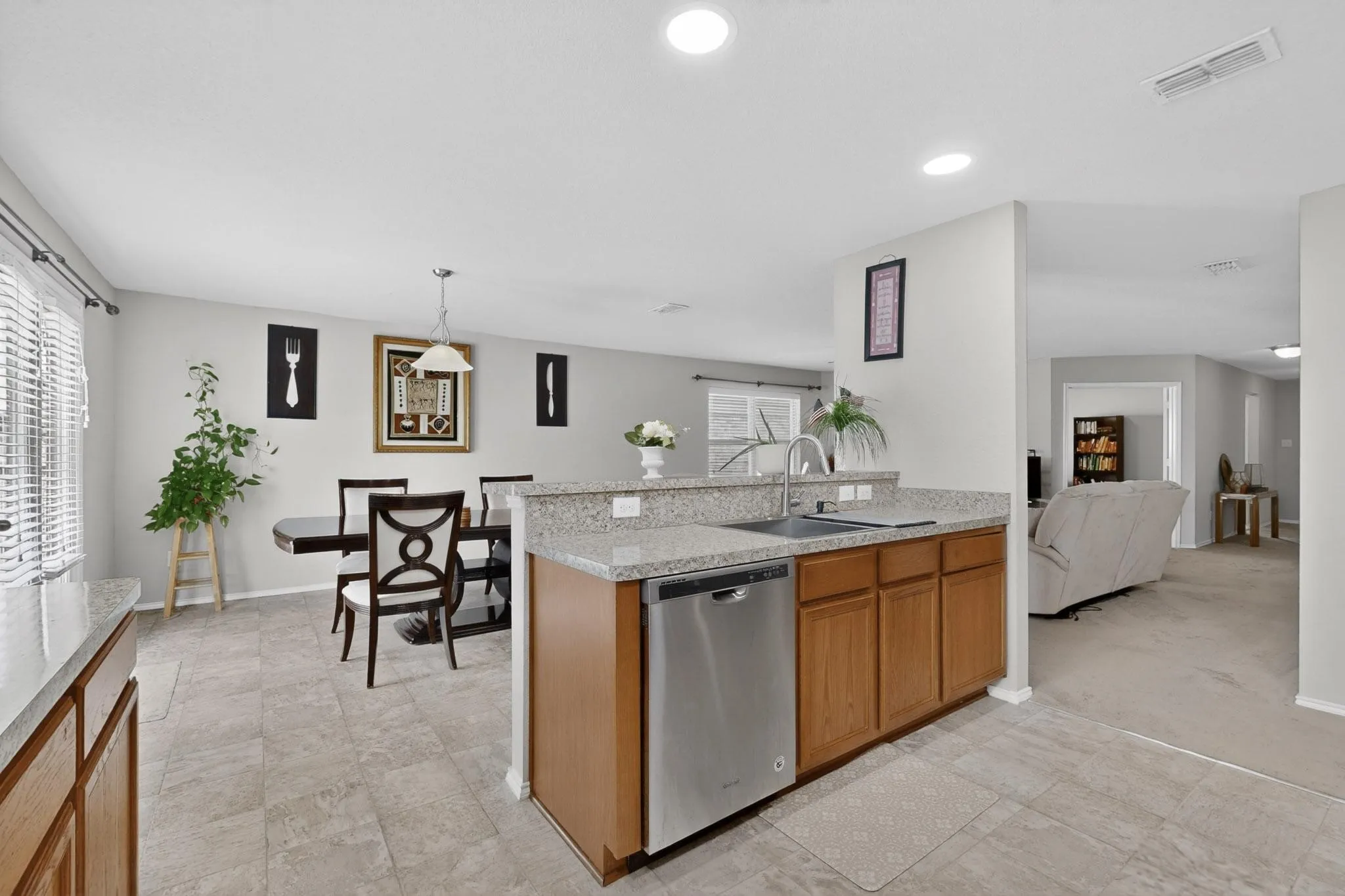 Kitchen with stainless steel dishwasher, wood finish cabinets, hanging light fixtures, light colored carpet, and light stone counters