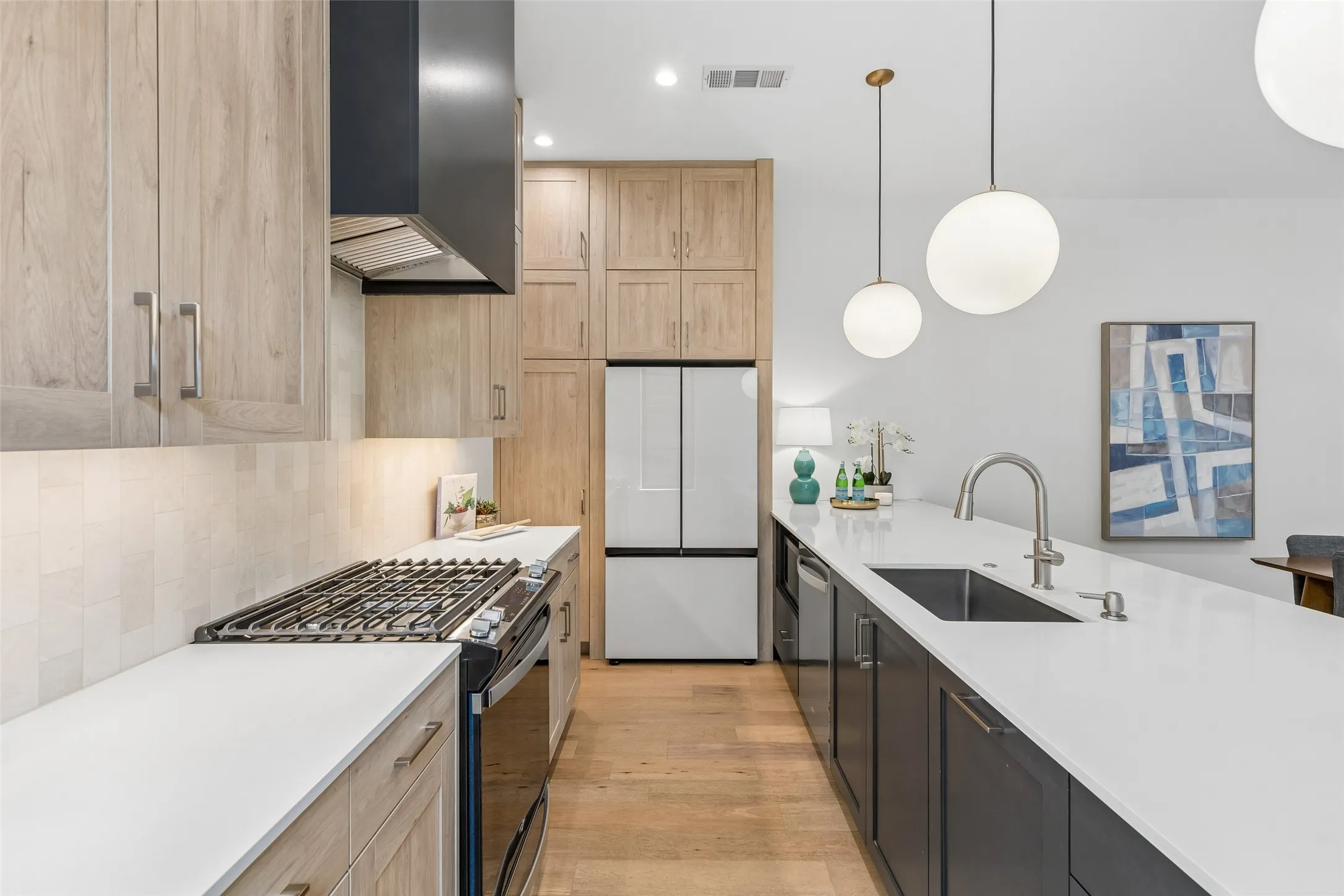 Two tone kitchen with dual tone cabinets, stainless steel appliances, light wood-style flooring, light stone countertops, and pendant lighting