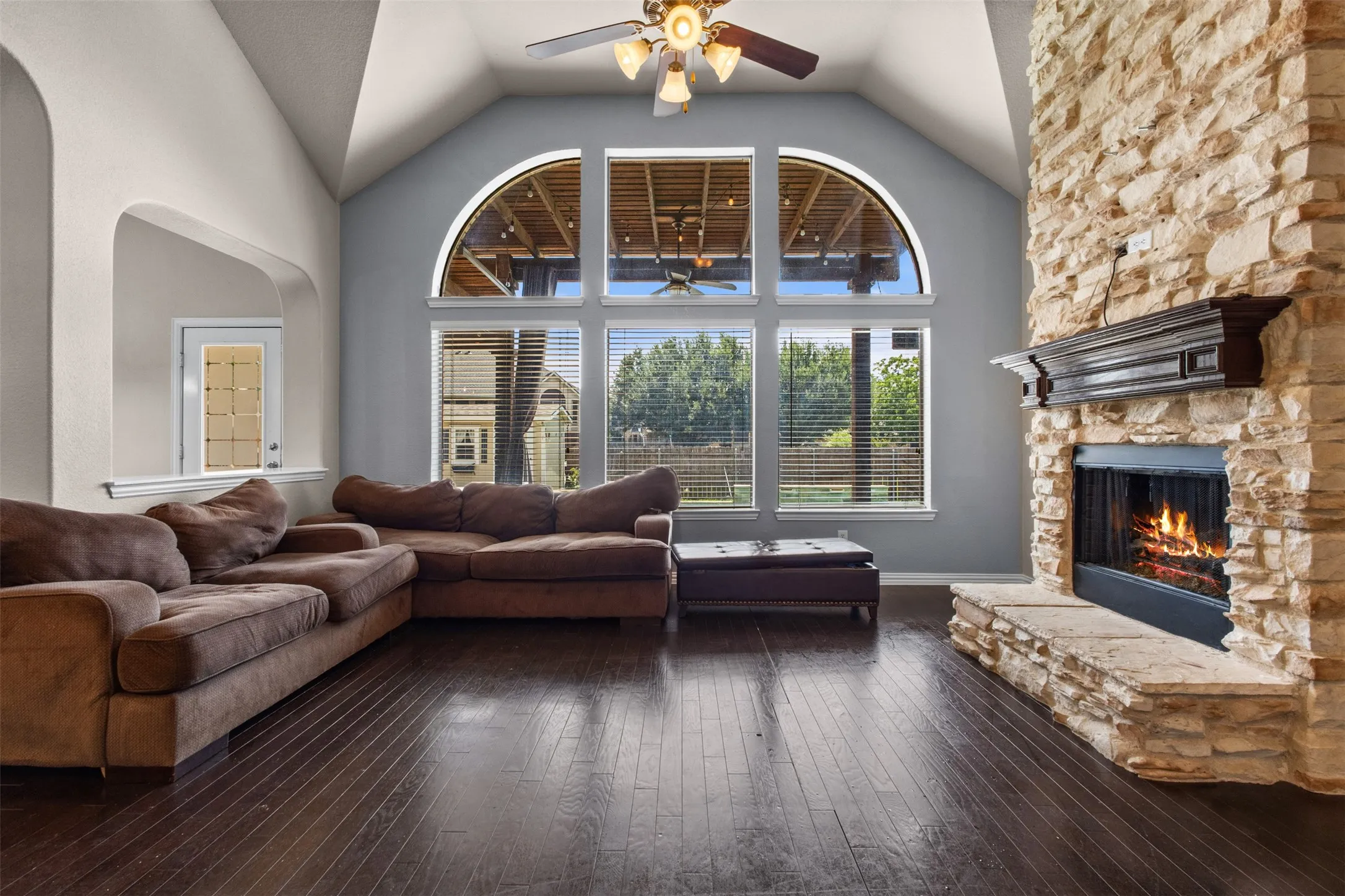 Living area featuring vaulted ceilings, wood-finish flooring, a stone-clad fireplace with a dark wood mantel, a multi-pane arched window, and a ceiling fan with integrated lighting