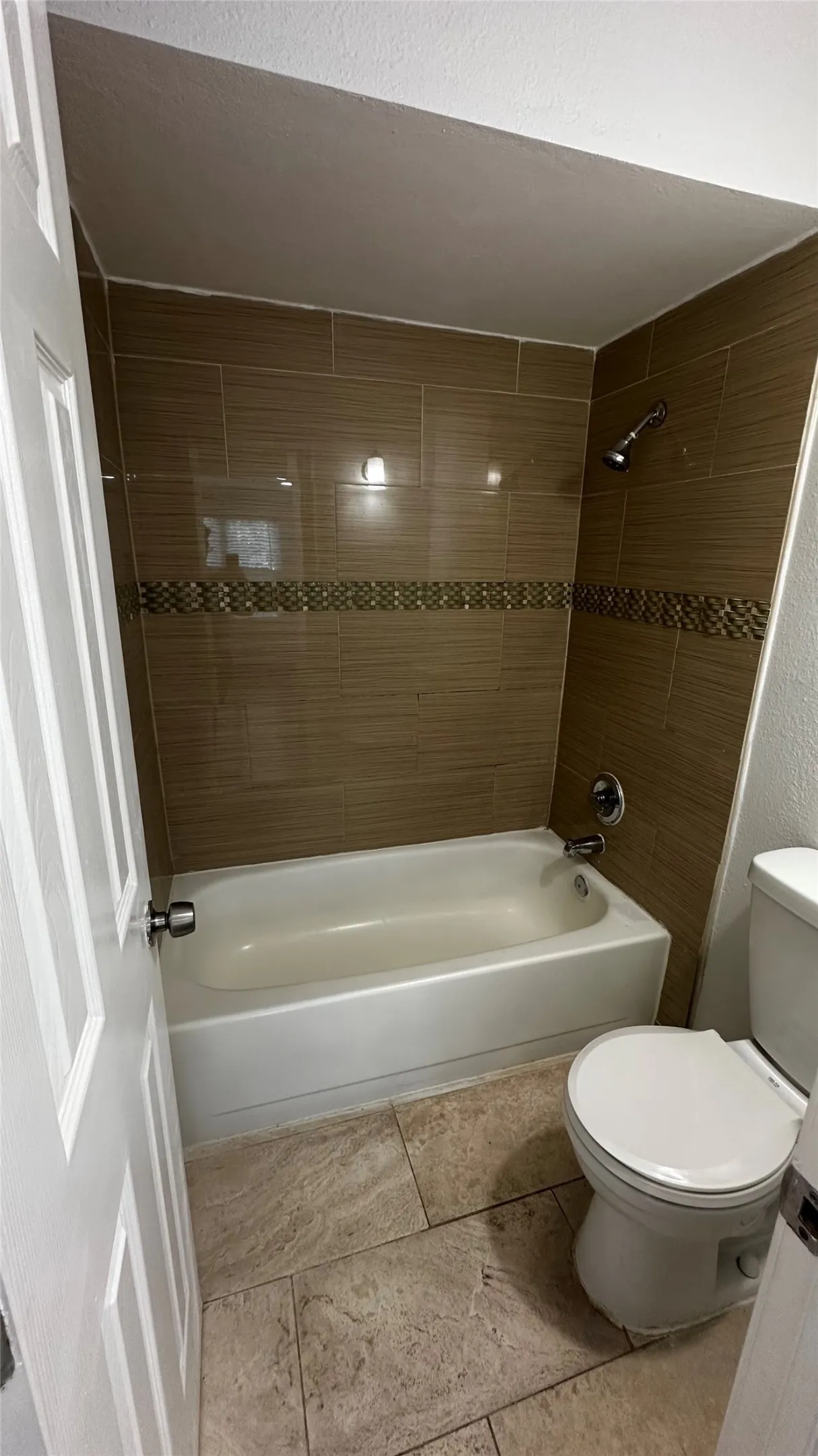 Bathroom featuring a white bathtub, ceramic tile shower surround with decorative accent band, shower head, toilet, and stone-look floor tiles
