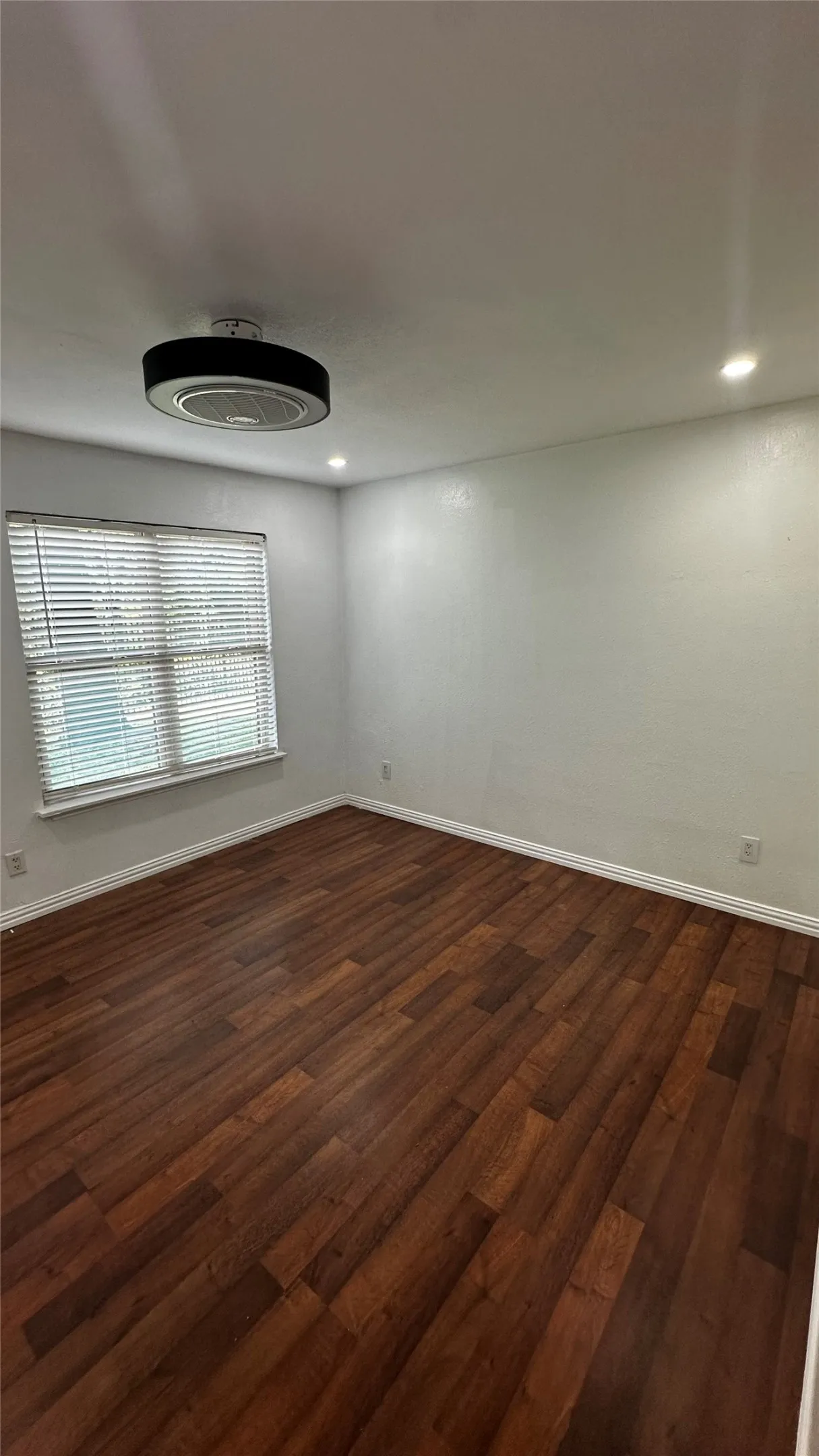 Spacious room featuring wood-finish flooring, white baseboards, and a window with blinds