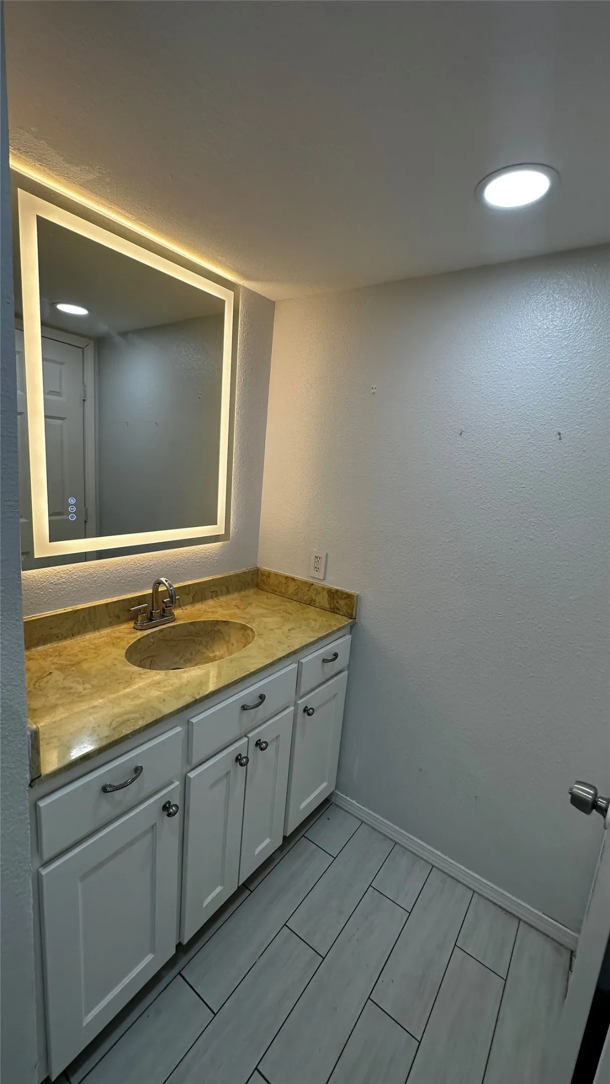 Vanity area featuring a luminous framed mirror, a granite-finish countertop, an undermount sink, white cabinetry with dark hardware, and wood-look tile flooring