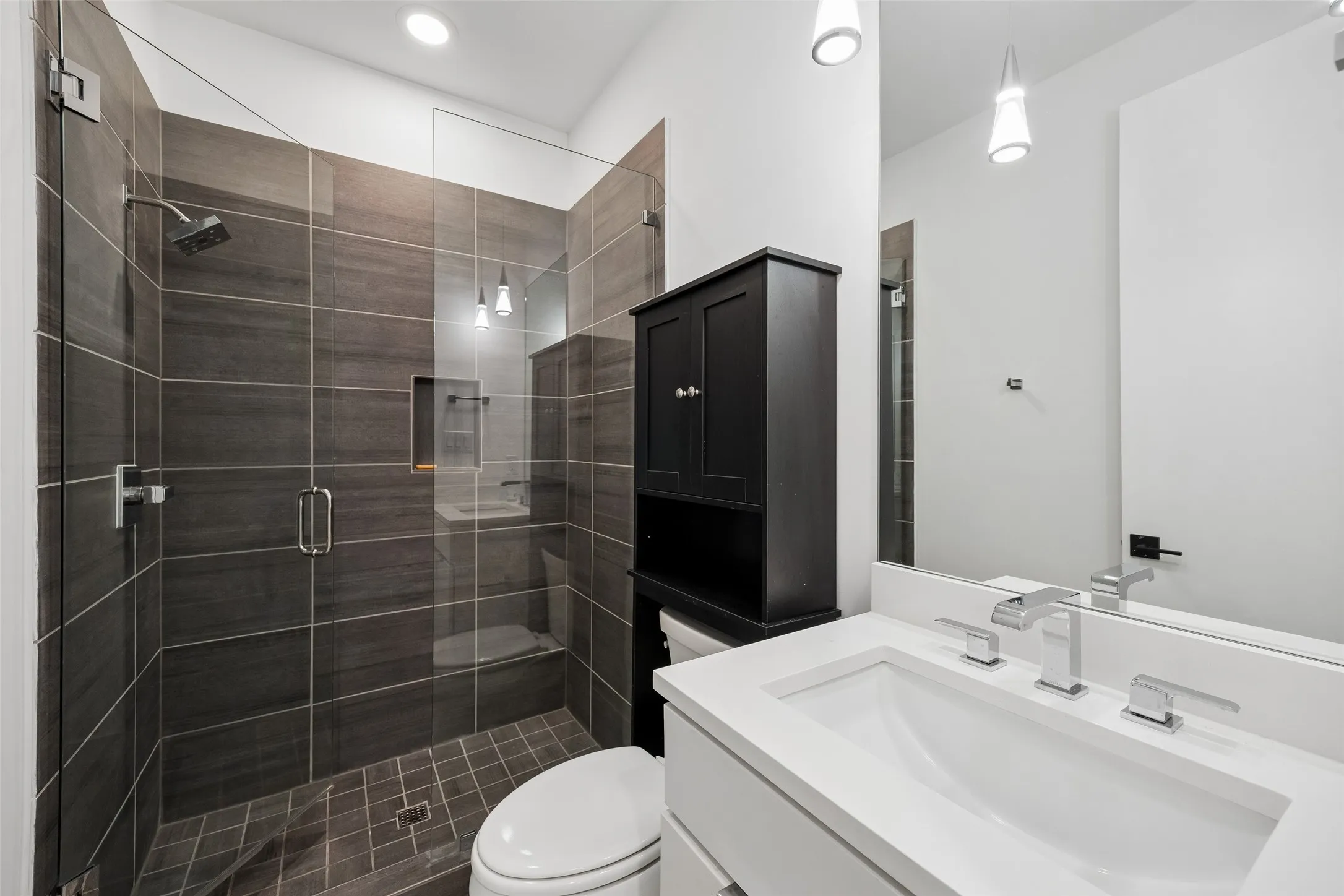 Modern bathroom featuring a vanity with a rectangular sink and contemporary fixtures, a spacious shower with dark tile surround and glass enclosure, and a built-in storage cabinet