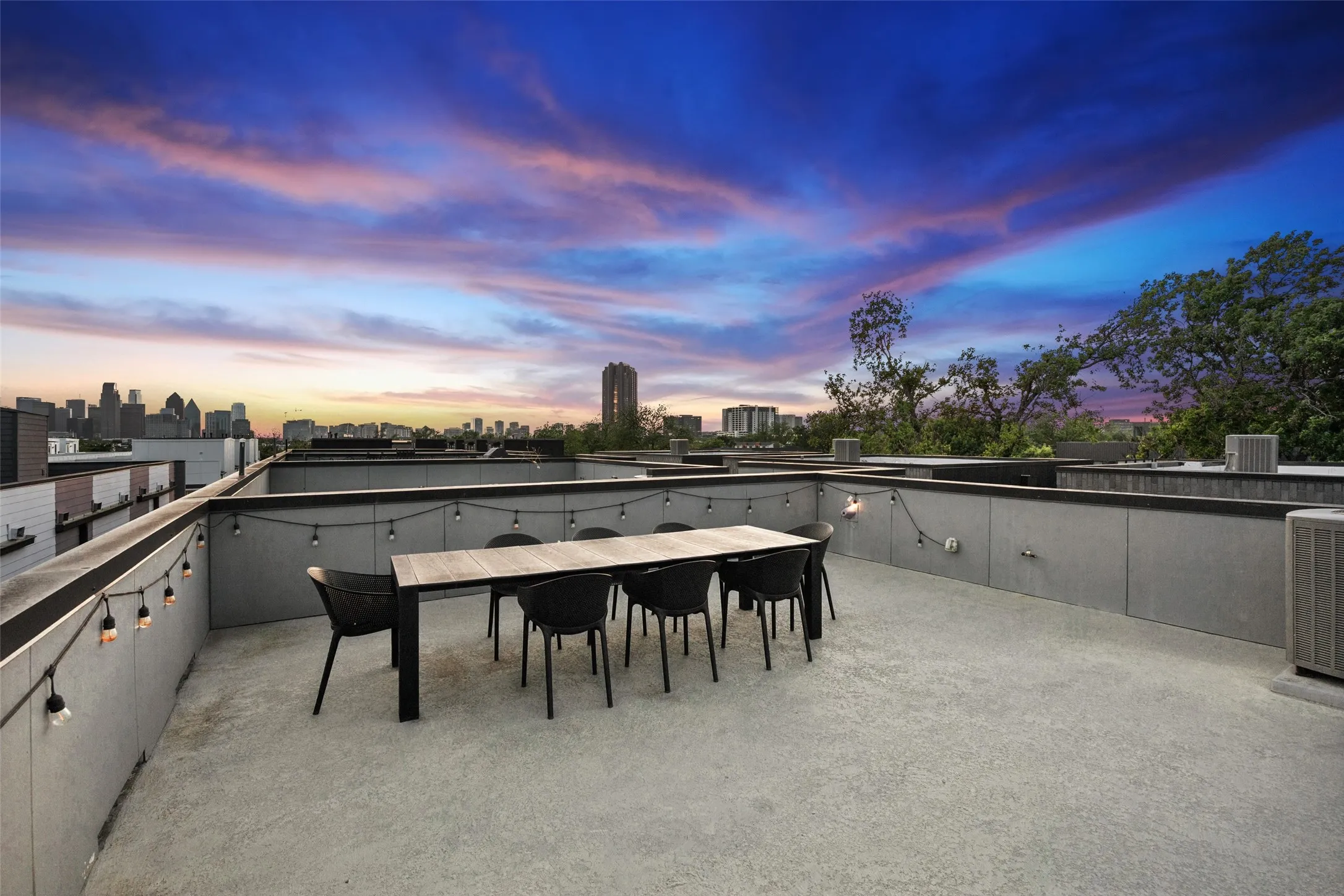 Expansive rooftop terrace featuring a concrete surface, perimeter wall, string lighting, and distant city buildings