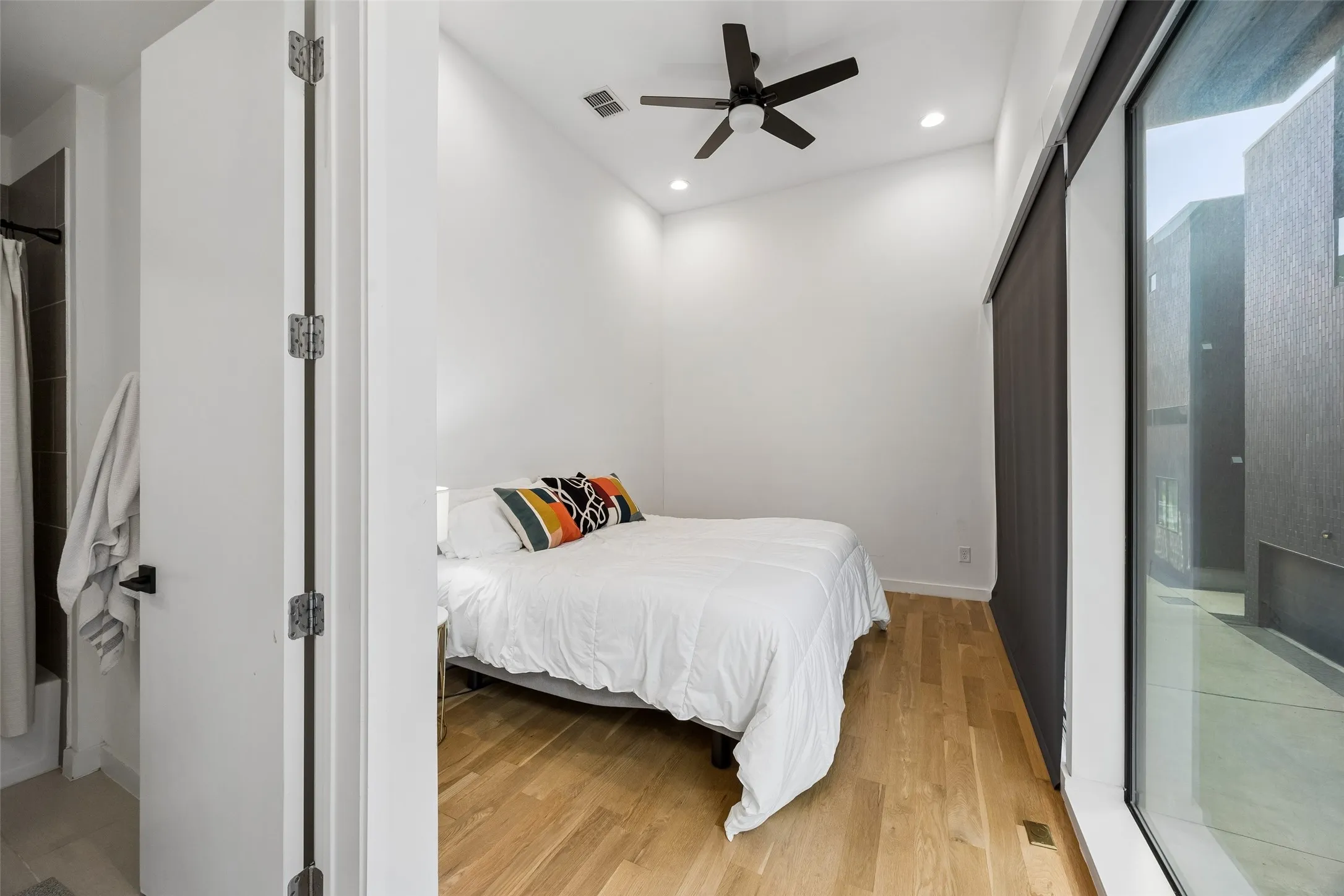 This bedroom features light wood flooring, a ceiling fan, recessed lighting, and a large window with a dark roller blind