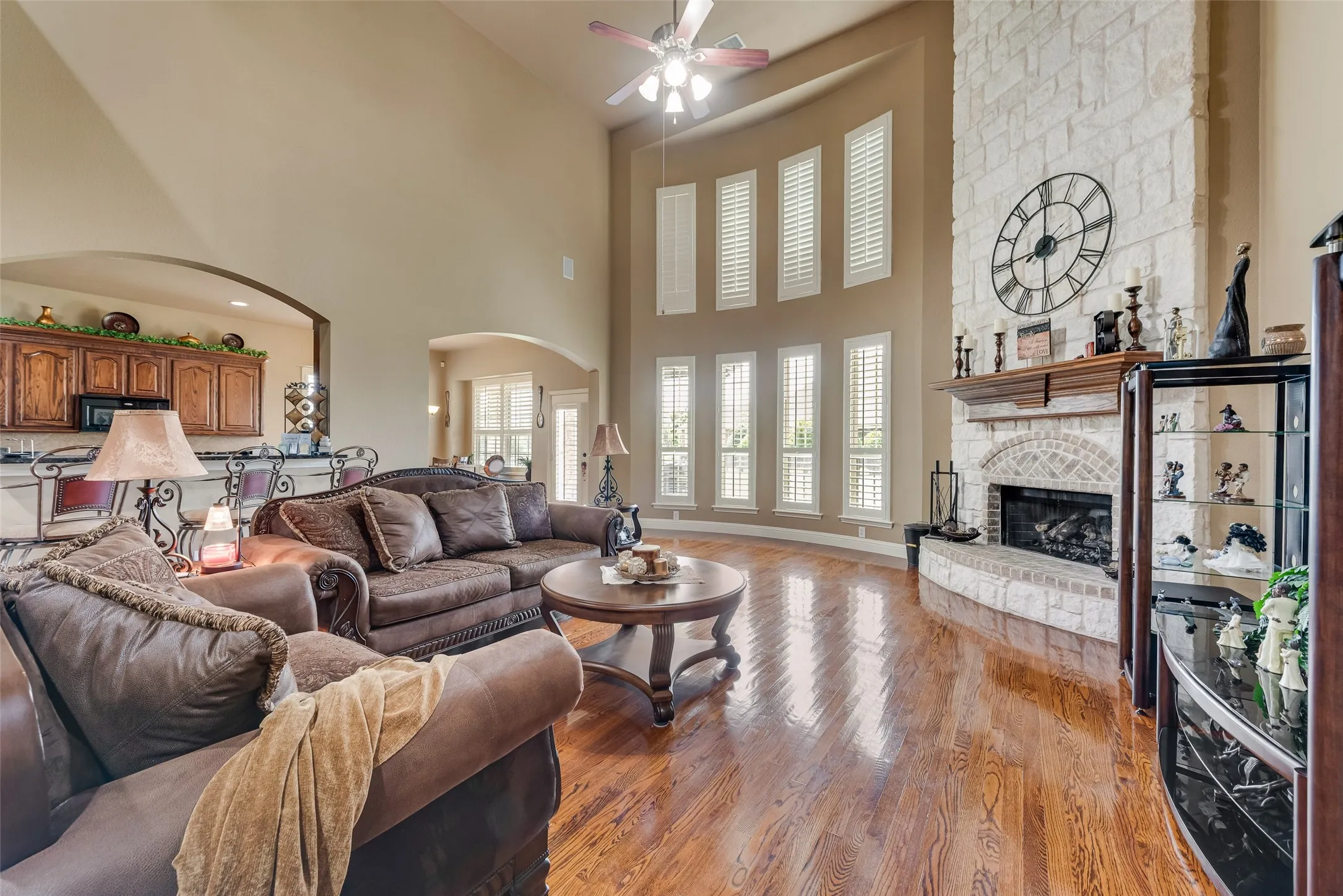 Living room with a stone fireplace, light wood finished floors, arched walkways, ceiling fan, and a high ceiling