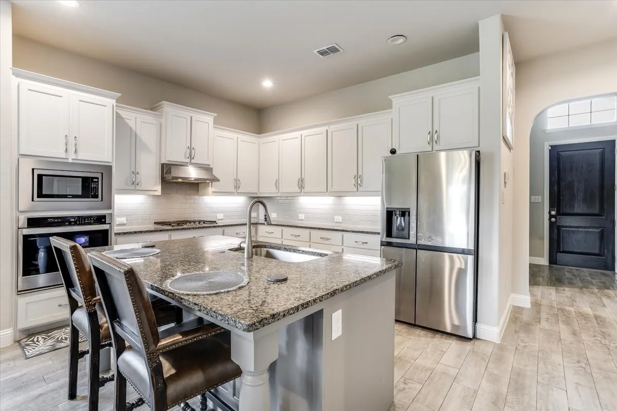 Kitchen featuring white cabinetry, a built-in oven and microwave, a stainless steel refrigerator, a large island with a sink, and wood-finish flooring