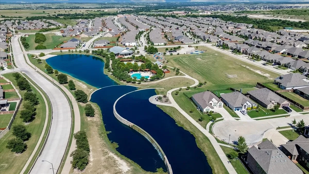 Aerial perspective of a developed community featuring multiple residential properties, a recreational pool area, a large open field, and a winding waterway