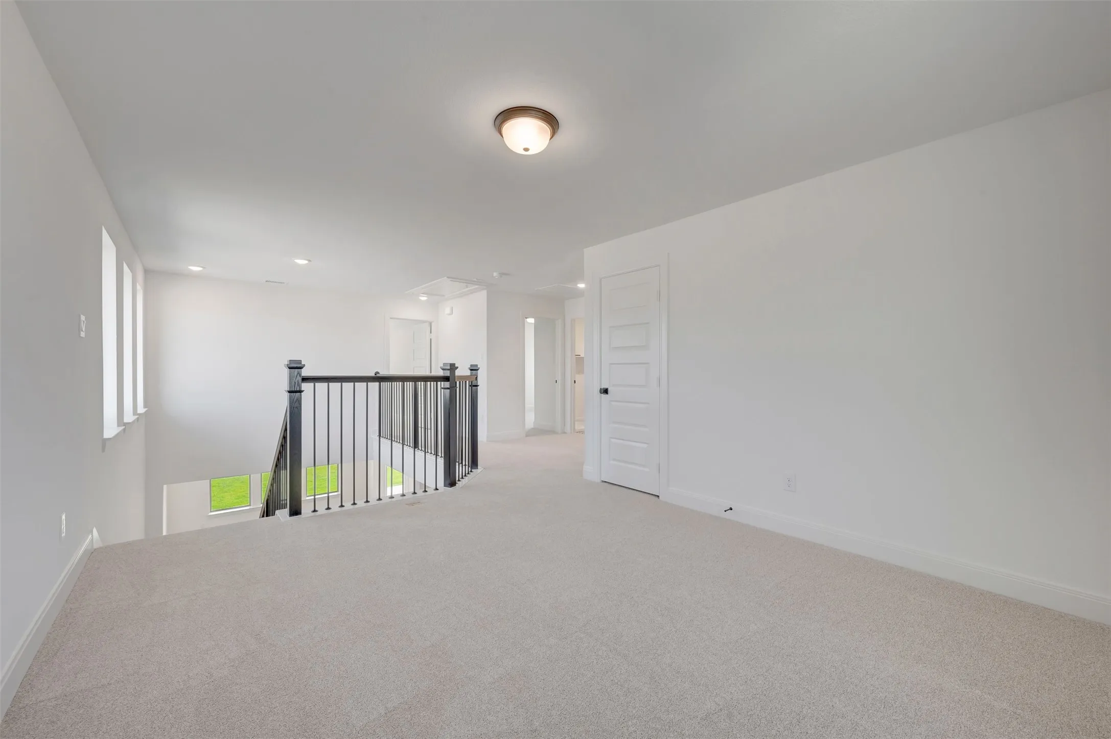 Spacious upper-level loft featuring light-colored carpeting, white walls, and a black iron and wood railing