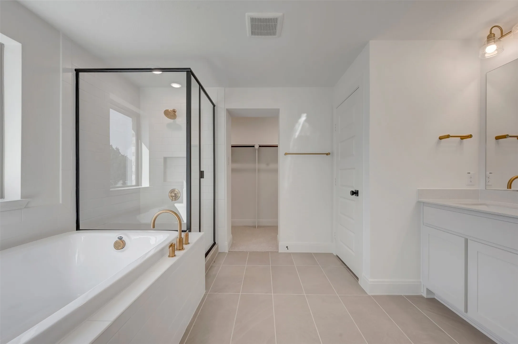Bathroom featuring a white soaking tub with brass fixtures, a glass-enclosed shower with black trim, a dual vanity with white cabinetry and countertops, and light-toned tile flooring