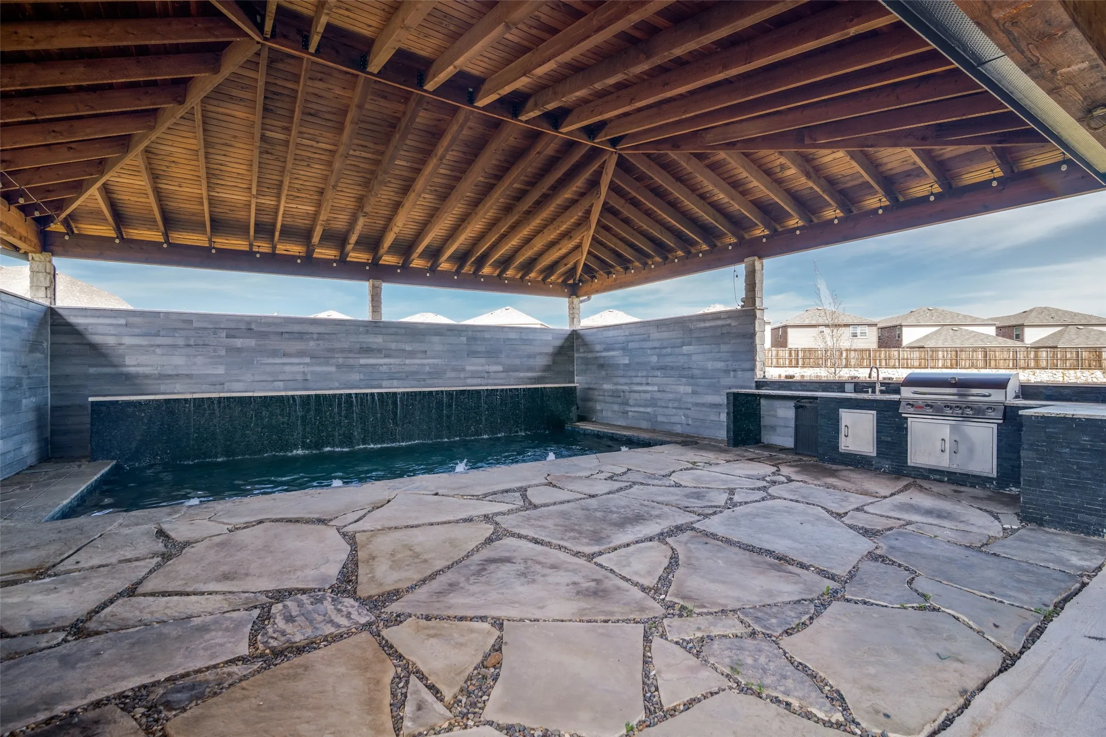Covered outdoor patio featuring a wood-beam ceiling, flagstone flooring, a built-in outdoor kitchen with a grill, and a water feature