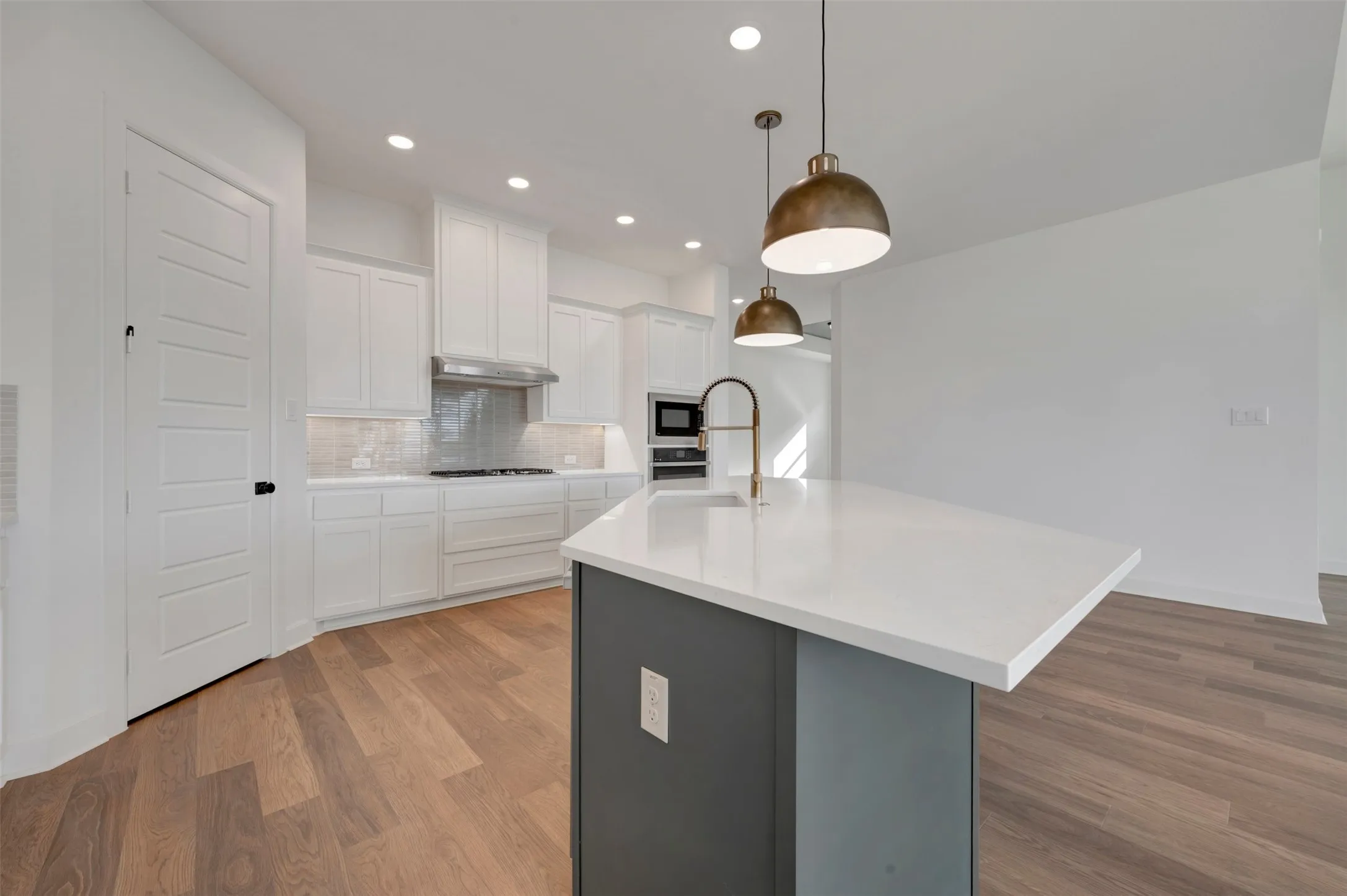 Modern kitchen featuring a central island with a white countertop and brass-finish faucet, under-cabinet lighting, stainless steel appliances, and wood-finish flooring