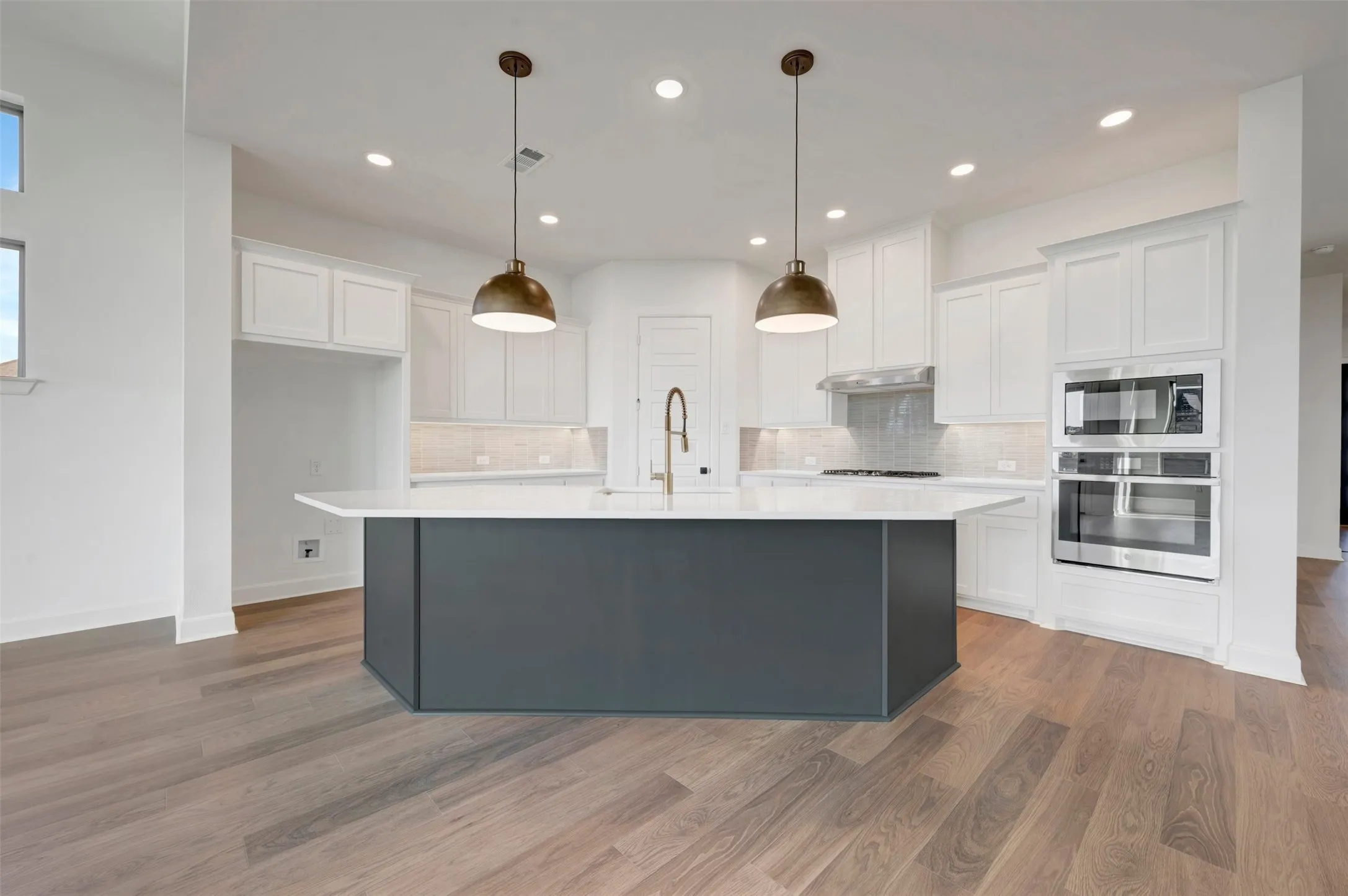 Kitchen featuring a dark-toned island with white countertop, brass pendant lighting, stainless steel built-in appliances, white cabinetry, and wood-finish flooring