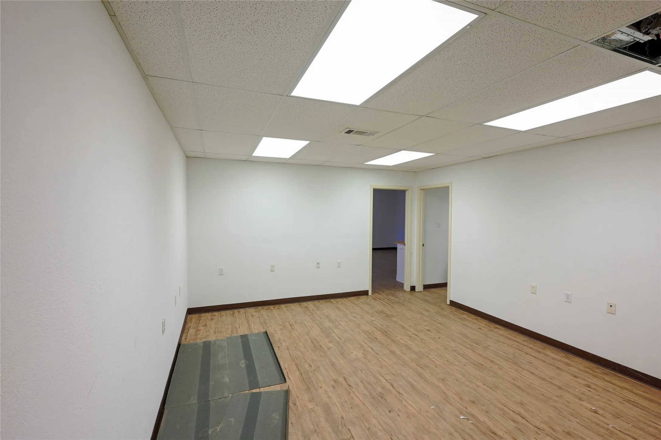 Spacious interior room with wood-finish flooring, white walls, and a dropped ceiling featuring recessed fluorescent lighting