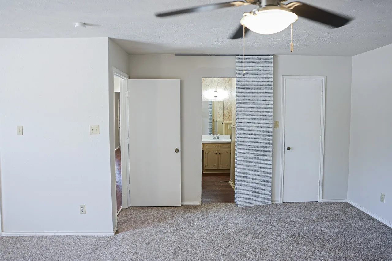 Carpeted room featuring a ceiling fan with integrated lighting, white walls, and a barn door entry to a bathroom with a wood-finish vanity