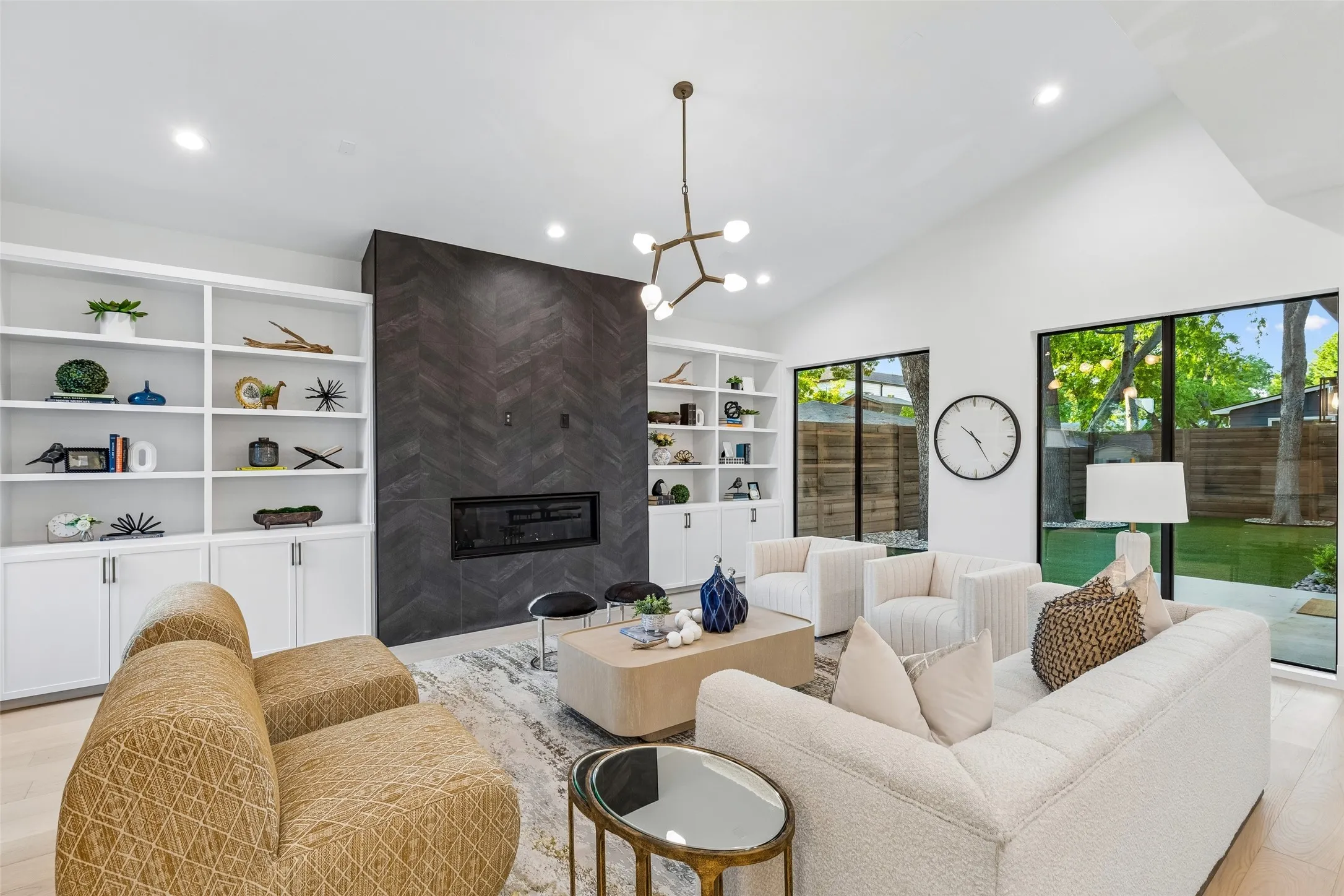 Open-concept living area with soaring ceilings, a sleek fireplace feature wall, and large windows that bring in natural light while offering views of the backyard, all flowing effortlessly into the kitchen.
