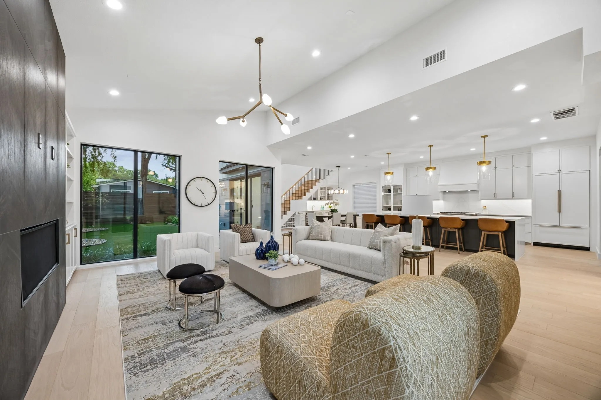 Light-filled living room with vaulted ceilings, statement fireplace wall, and expansive windows overlooking the backyard, all opening seamlessly to the kitchen for effortless entertaining.