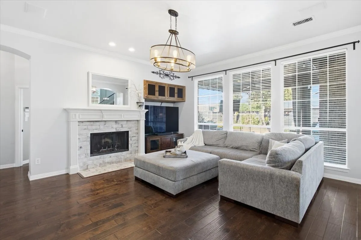 Dark wood-finish flooring, a white brick fireplace with a mantel, multiple windows with blinds, recessed lighting, and a decorative overhead light fixture