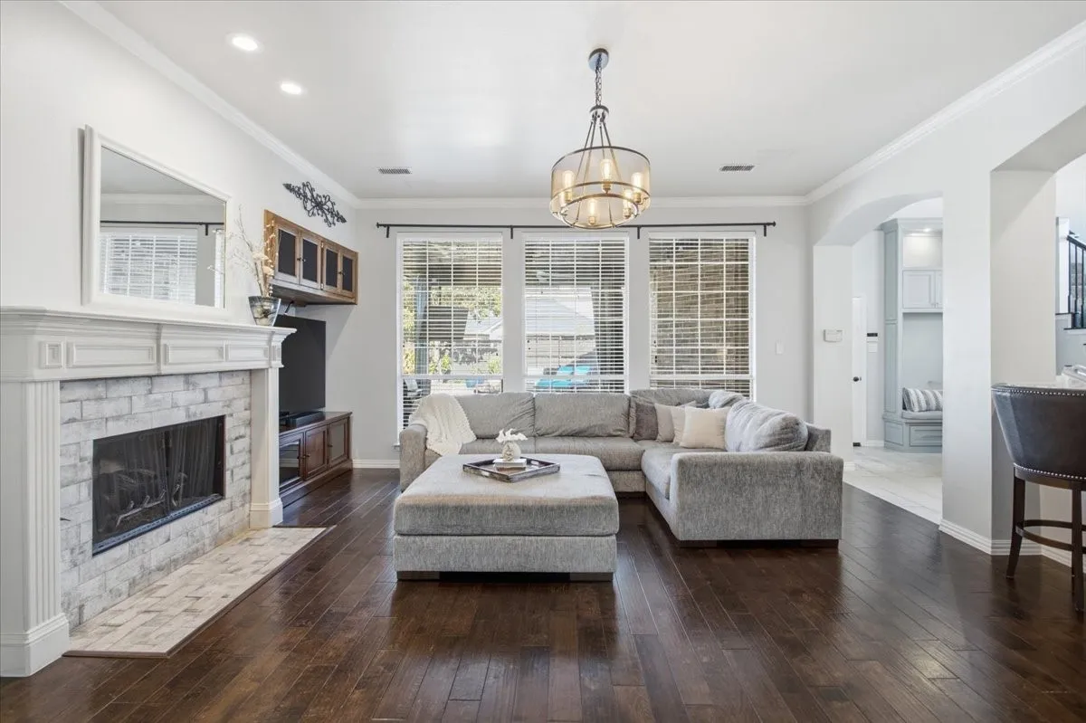 Dark wood-finish flooring, white brick fireplace with mantel, crown molding, built-in media cabinet, and a multi-tier chandelier