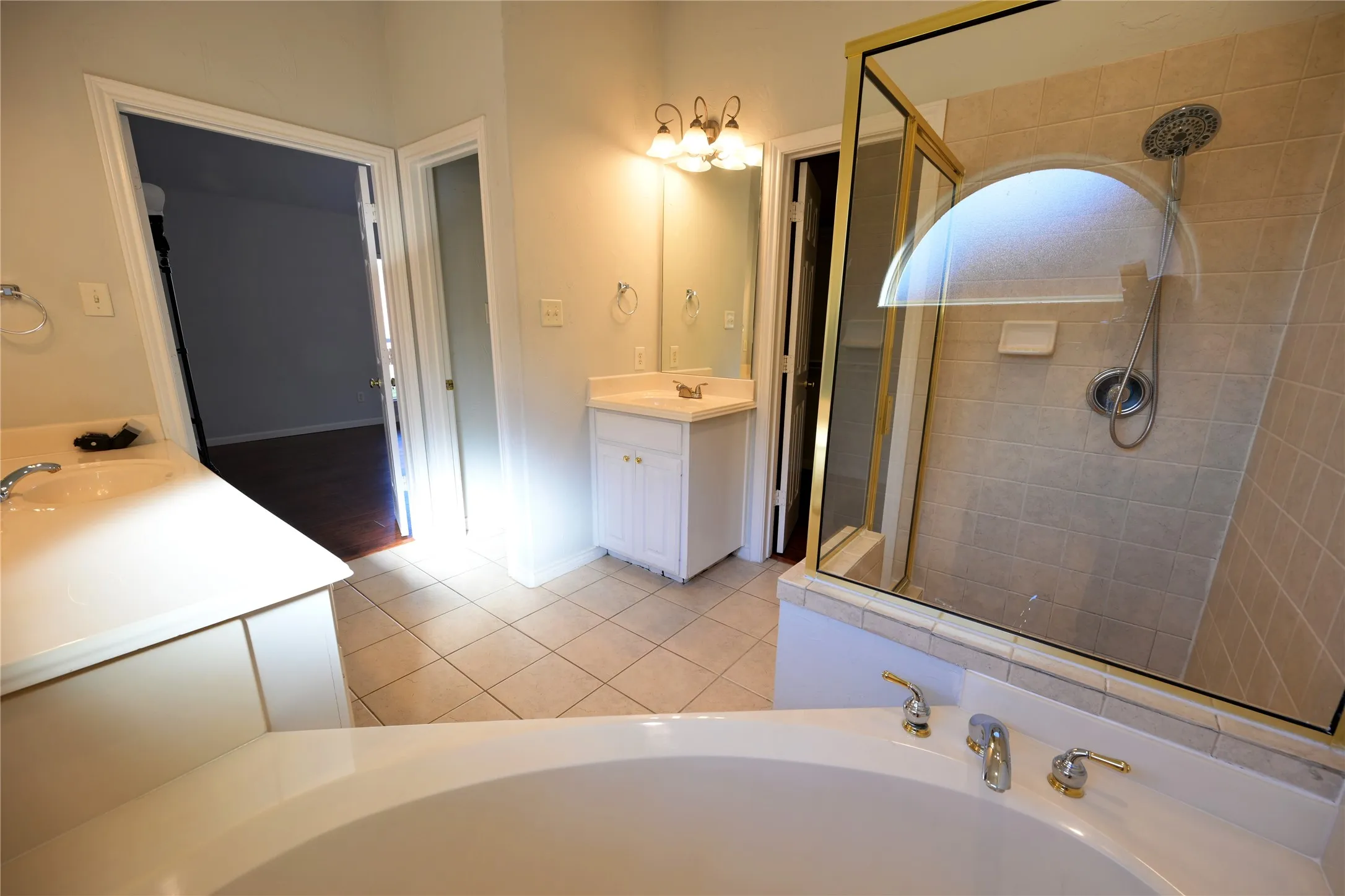 Bathroom featuring a corner soaking tub with deck-mounted fixtures, separate shower with tile surround and glass enclosure, tile flooring, and dual vanity areas with white cabinetry and integrated sinks