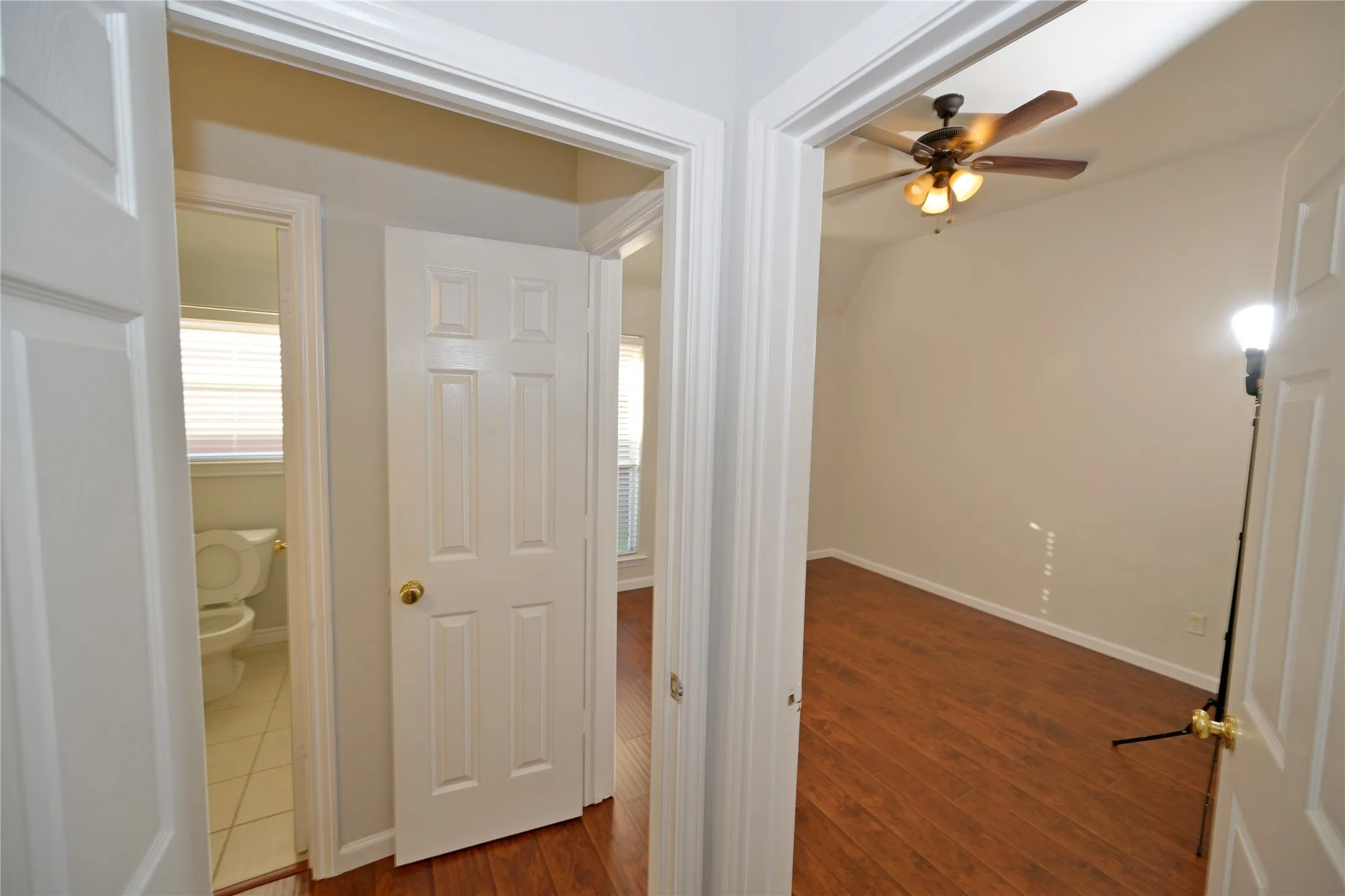 Interior hallway with white paneled doors leading to a room with wood-finish flooring and a ceiling fan, and a bathroom with tile flooring and a toilet
