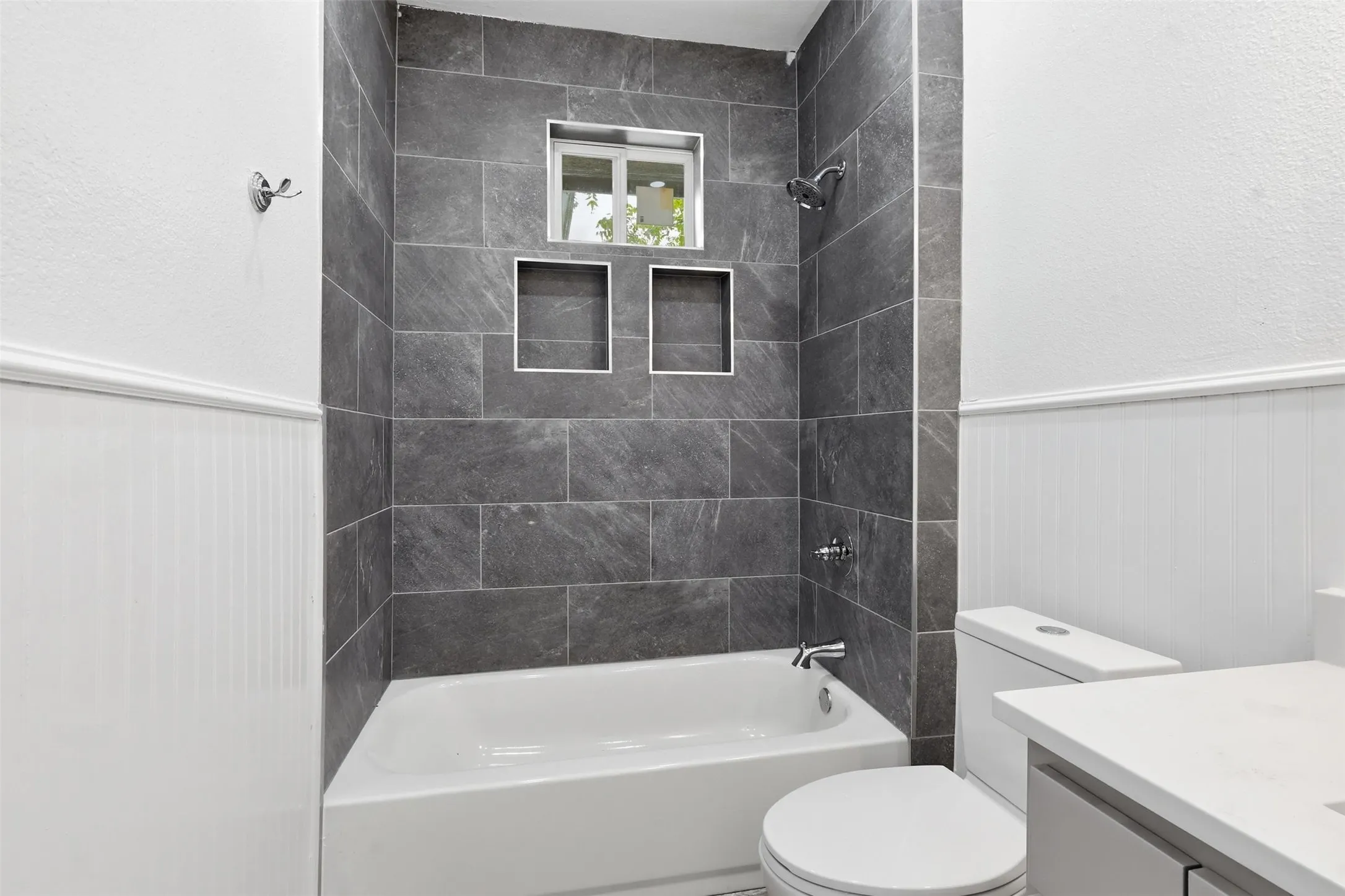 Bathtub with dark gray stone-look tile surround featuring a built-in window and two tiled niches