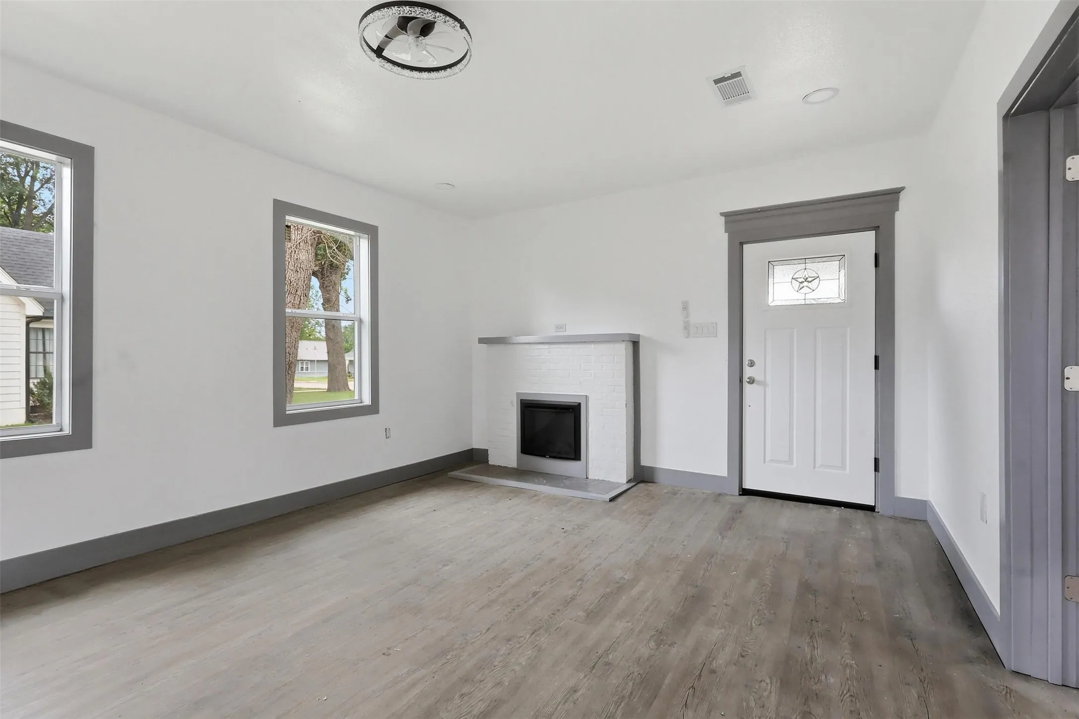 Inviting living space featuring wood-finish flooring, two large windows with gray trim, a decorative fireplace with a tile surround, a white entry door with a decorative glass insert, and a modern ceiling fixture