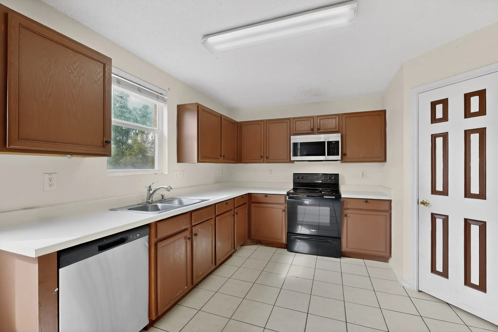Kitchen with black range with electric stovetop, light countertops, stainless steel dishwasher, wood finish cabinets, and light tile patterned floors