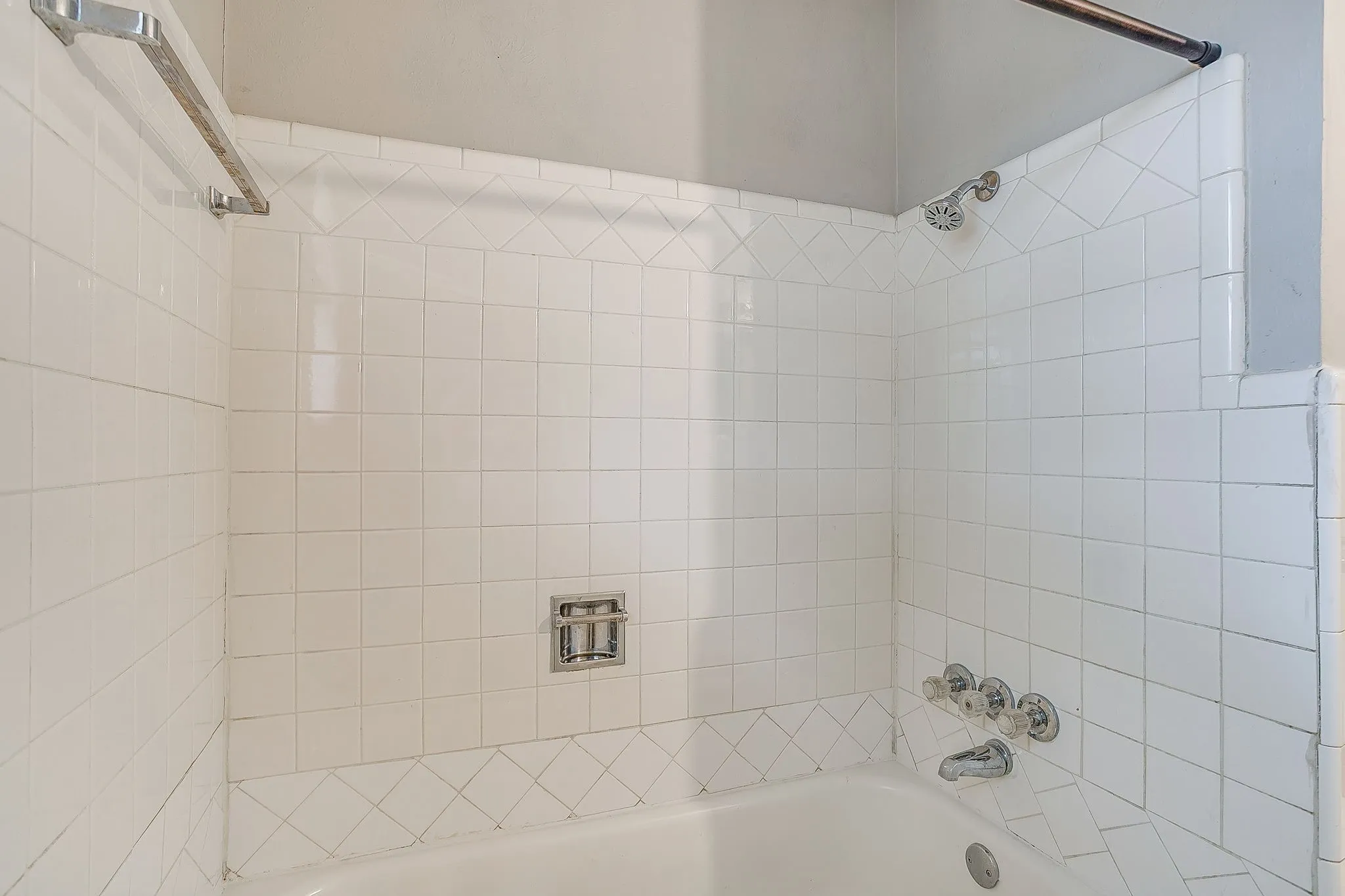 Bathtub and shower combination featuring white square tiles, decorative tile banding, and chrome fixtures