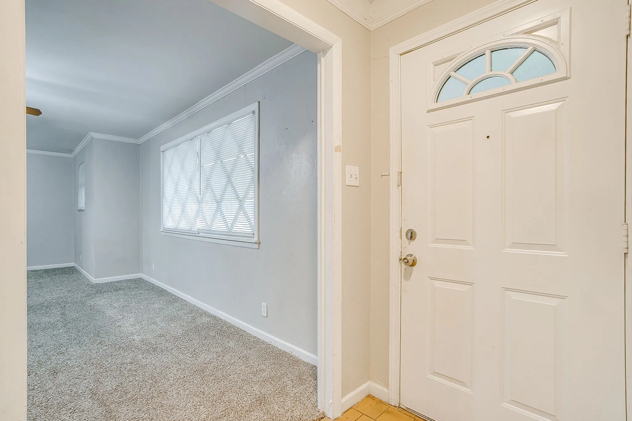 Entryway featuring a paneled door with an arched transom window, ceramic tile flooring, and direct access to a carpeted room with crown molding and window blinds