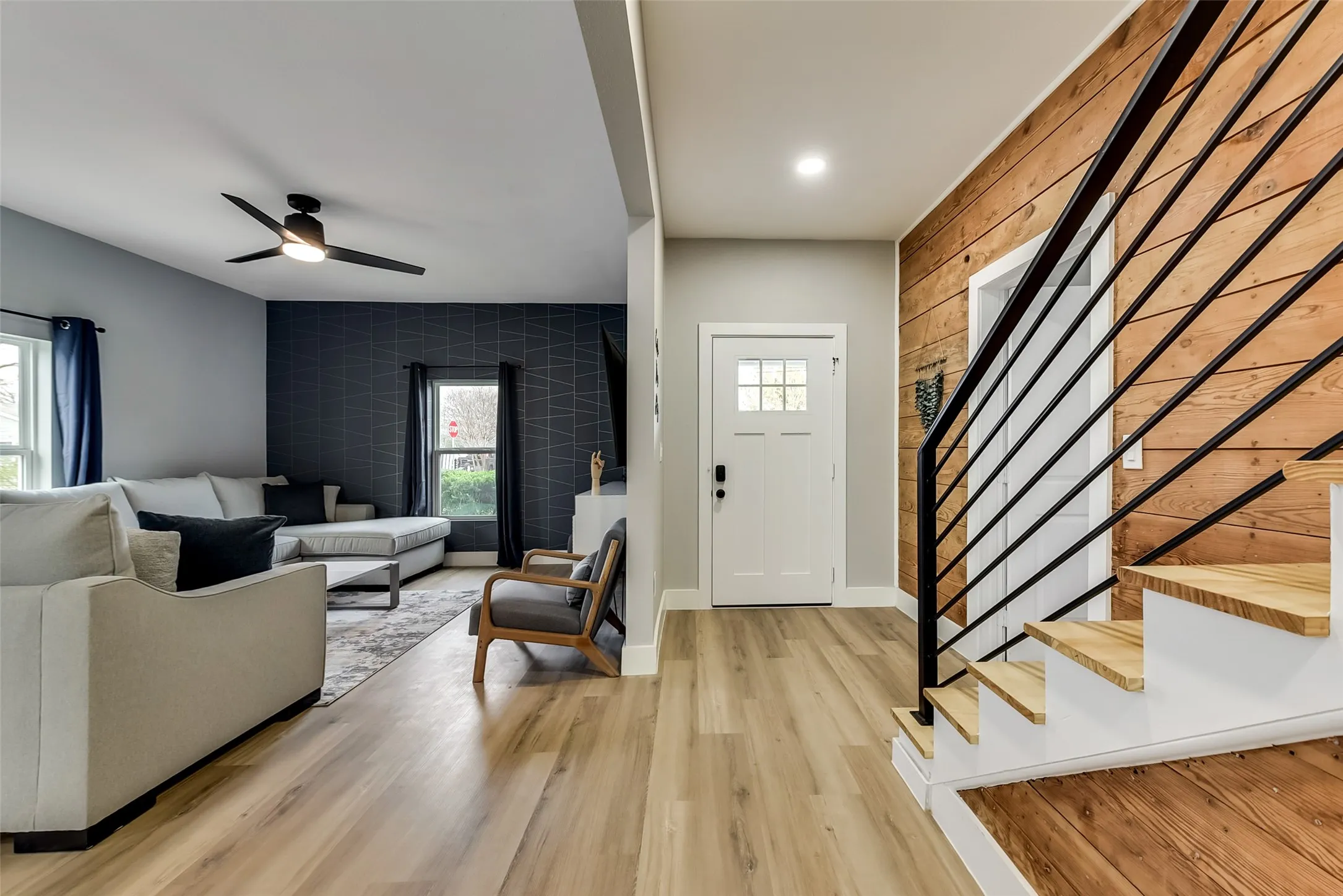 Living area with a ceiling fan, baseboards, light wood finished floors, an accent wall, and stairway