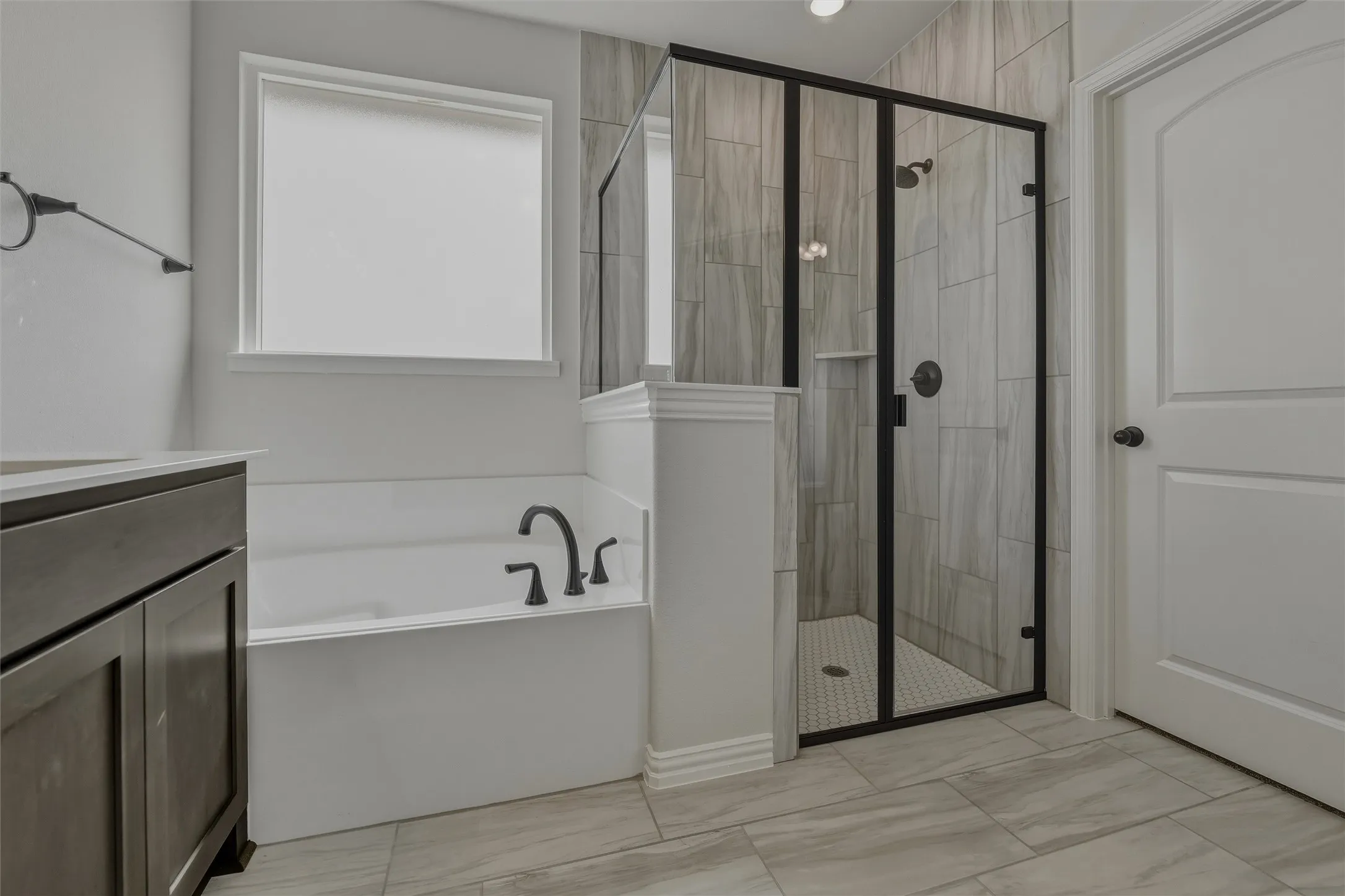 Frameless glass shower enclosure with matte black hardware, white soaking tub with matte black fixtures, wood-finish vanity with countertop, and large format tile flooring