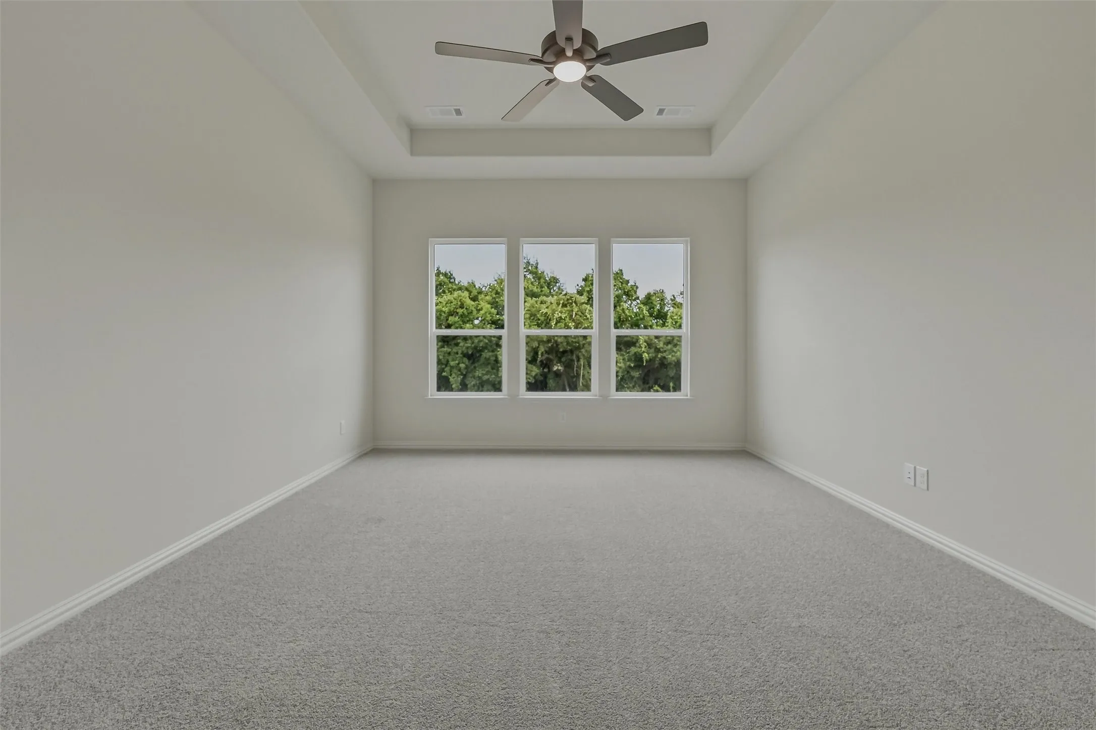 Room featuring three large windows, a tray ceiling, a ceiling fan with integrated lighting, neutral wall paint, and grey carpeting