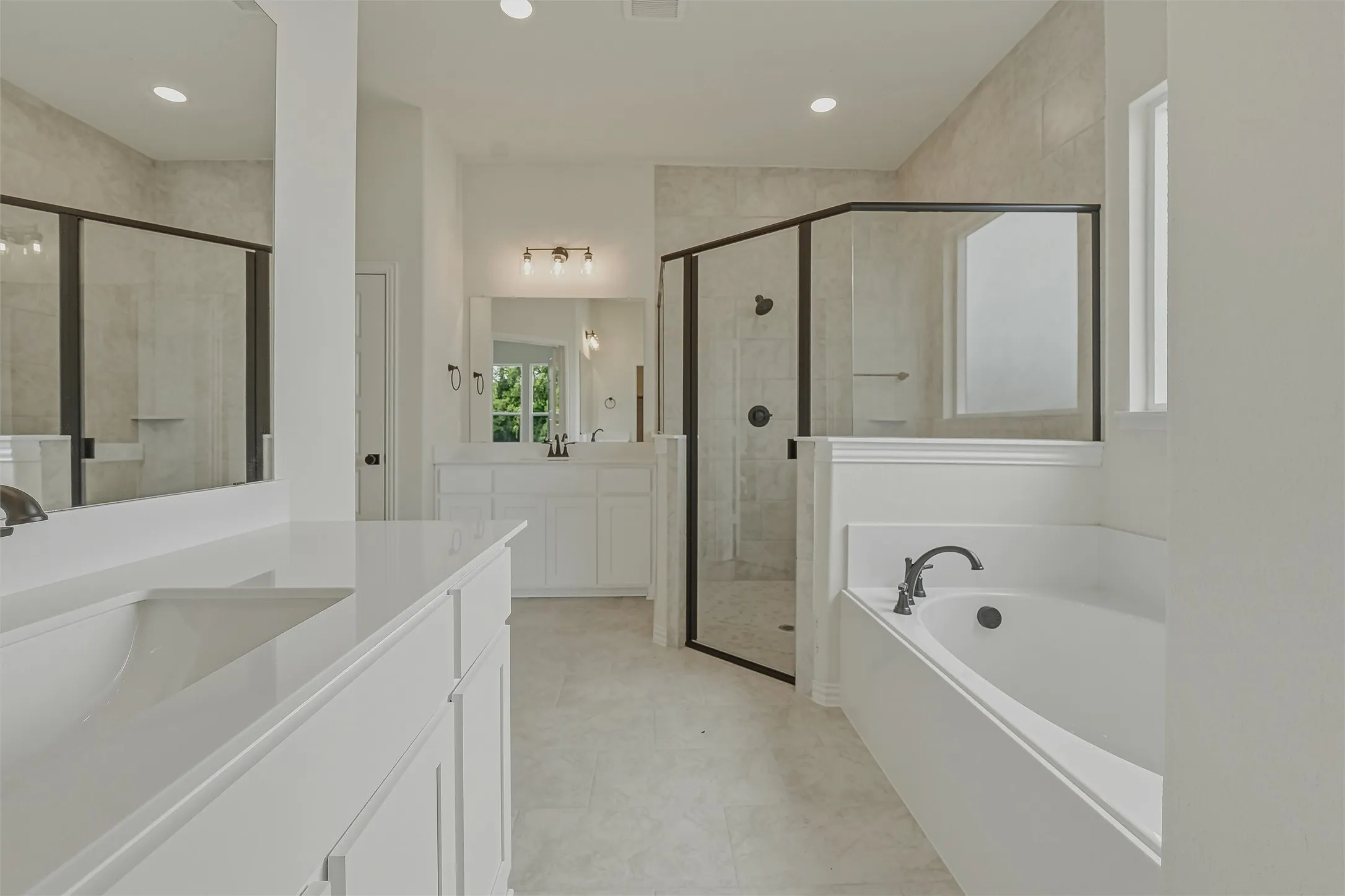 Spacious bathroom featuring a built-in soaking tub, a separate glass-enclosed shower, dual vanities with integrated sinks, white cabinetry, and recessed lighting
