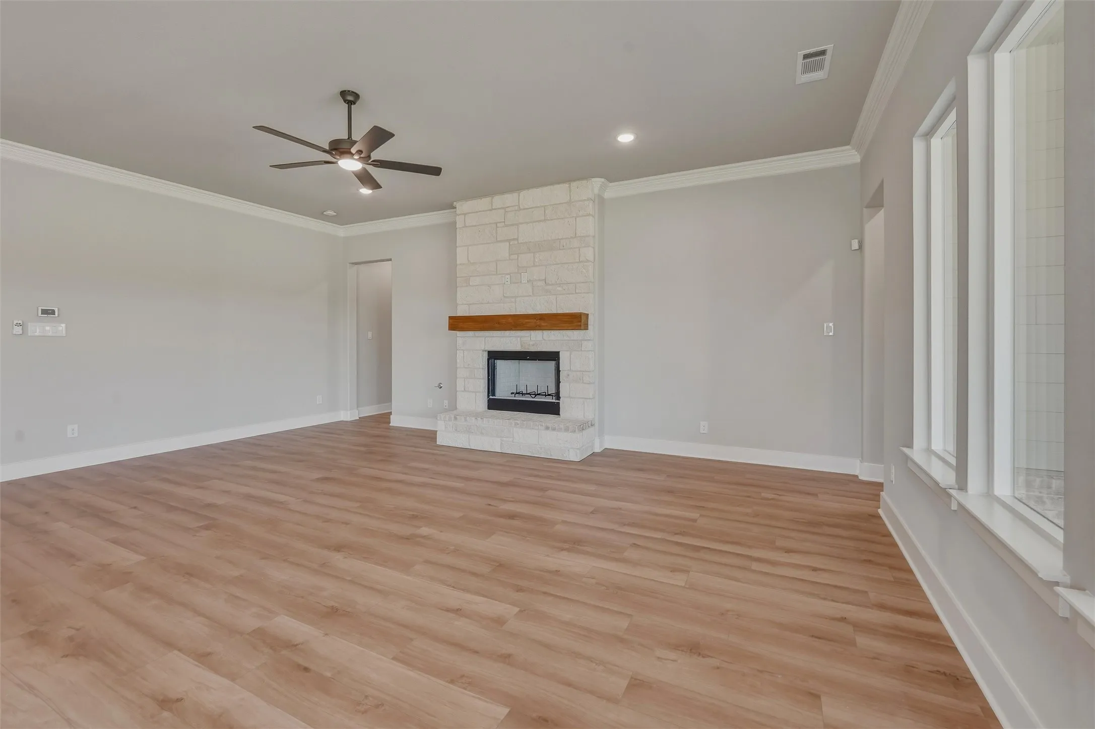 Spacious living area featuring light wood-finish flooring, a stone fireplace with a wood mantel, crown molding, and multiple windows