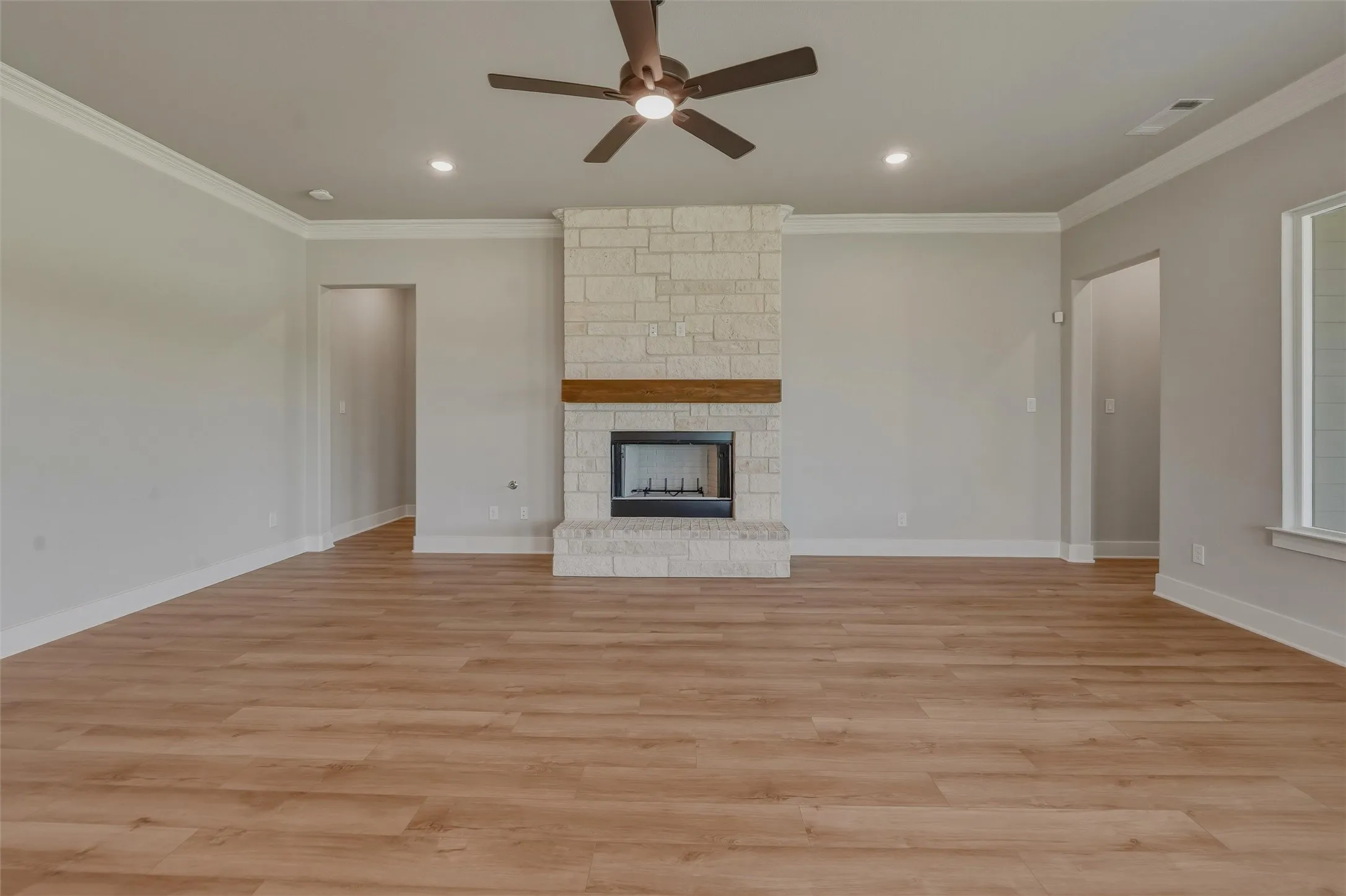 Spacious living area featuring wood-finish flooring, a stone-clad fireplace with a wood mantel, crown molding, a ceiling fan, and recessed lighting