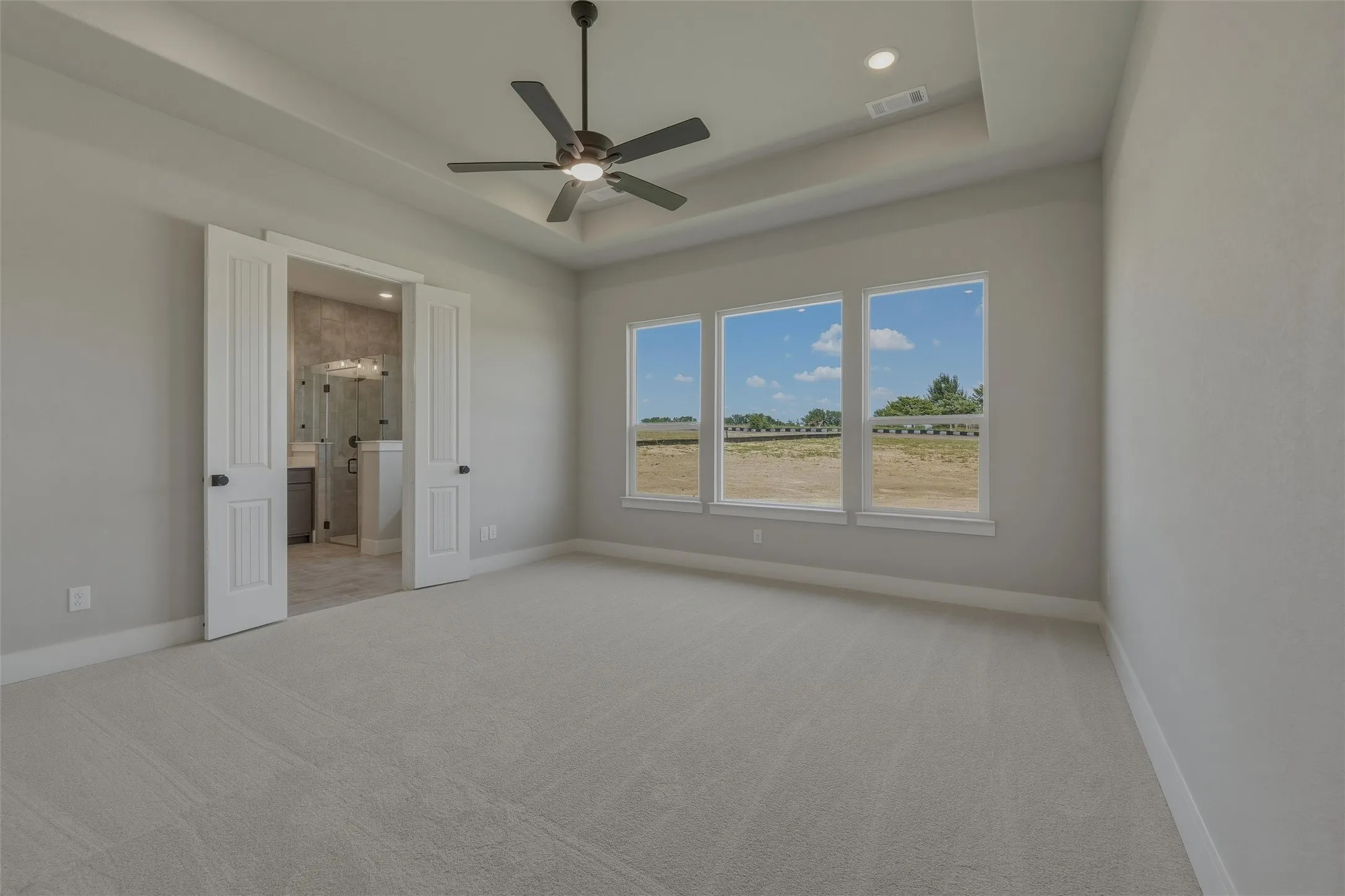 Spacious room featuring light-colored carpeting, a tray ceiling with a ceiling fan, and three large windows