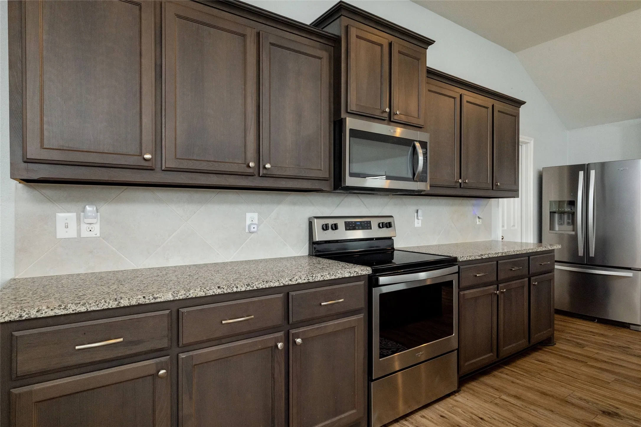 Kitchen with stainless steel appliances, dark wood finish cabinetry, light stone countertops, lofted ceiling, and dark wood finished floors