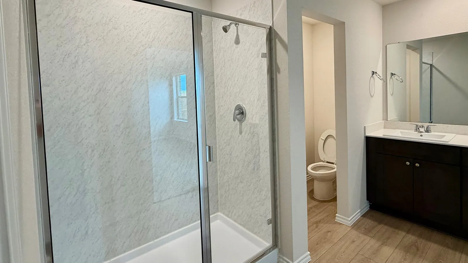 Shower enclosure with a clear glass door and a white patterned surround, wood-finish flooring, a dark wood-finish vanity with a light-colored countertop, and a rectangular mirror