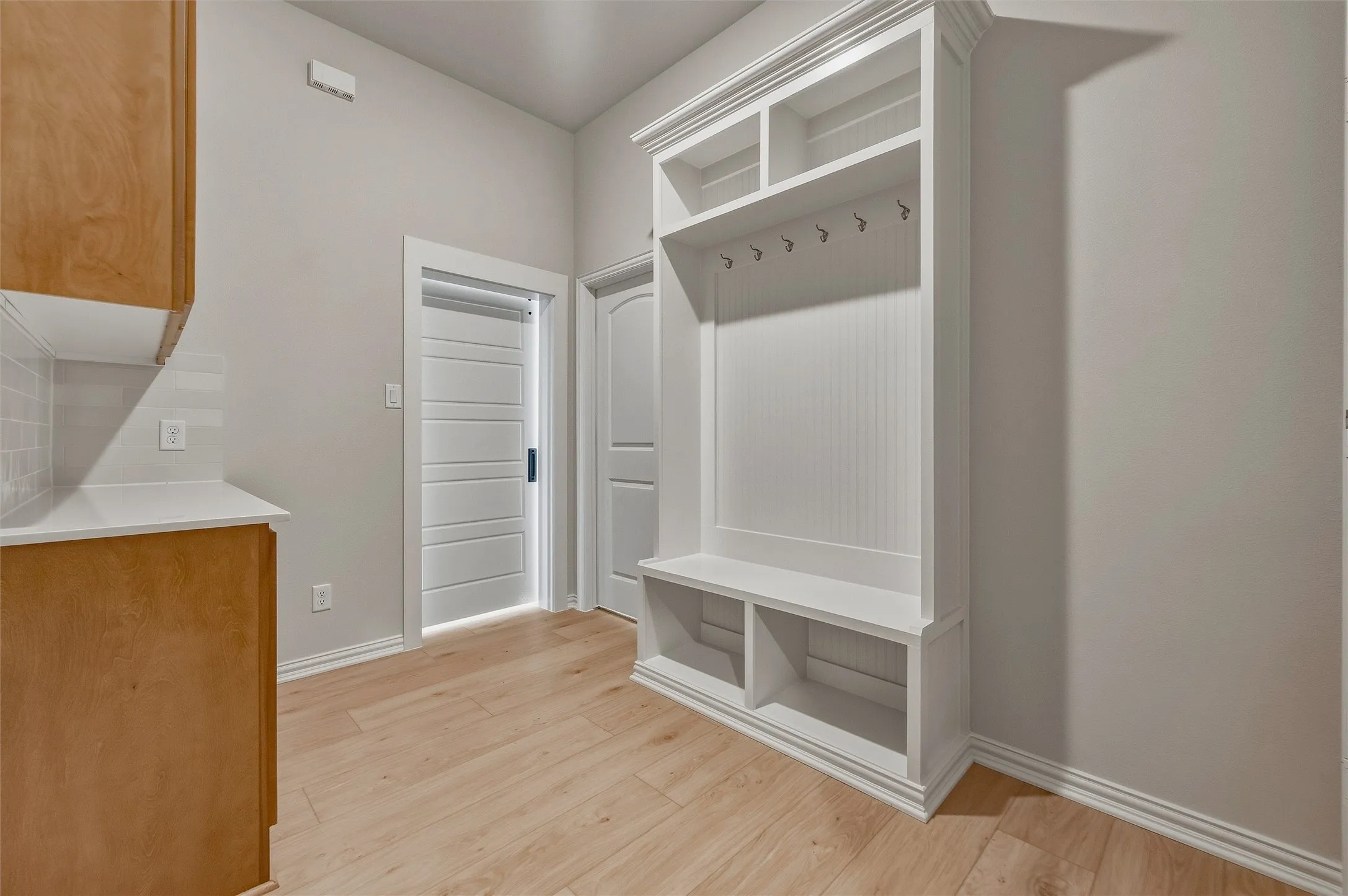 Built-in white storage unit with cubbies and hooks, wood-finish flooring, light-toned cabinetry, white countertop, and subway tile backsplash