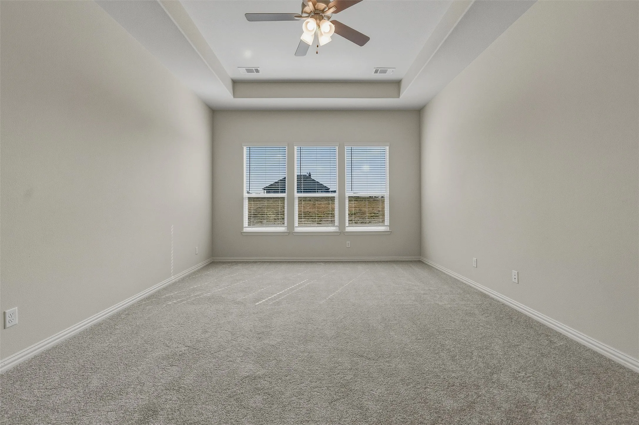 Tray ceiling with a ceiling fan, plush gray carpeting, and a three-panel window with horizontal blinds