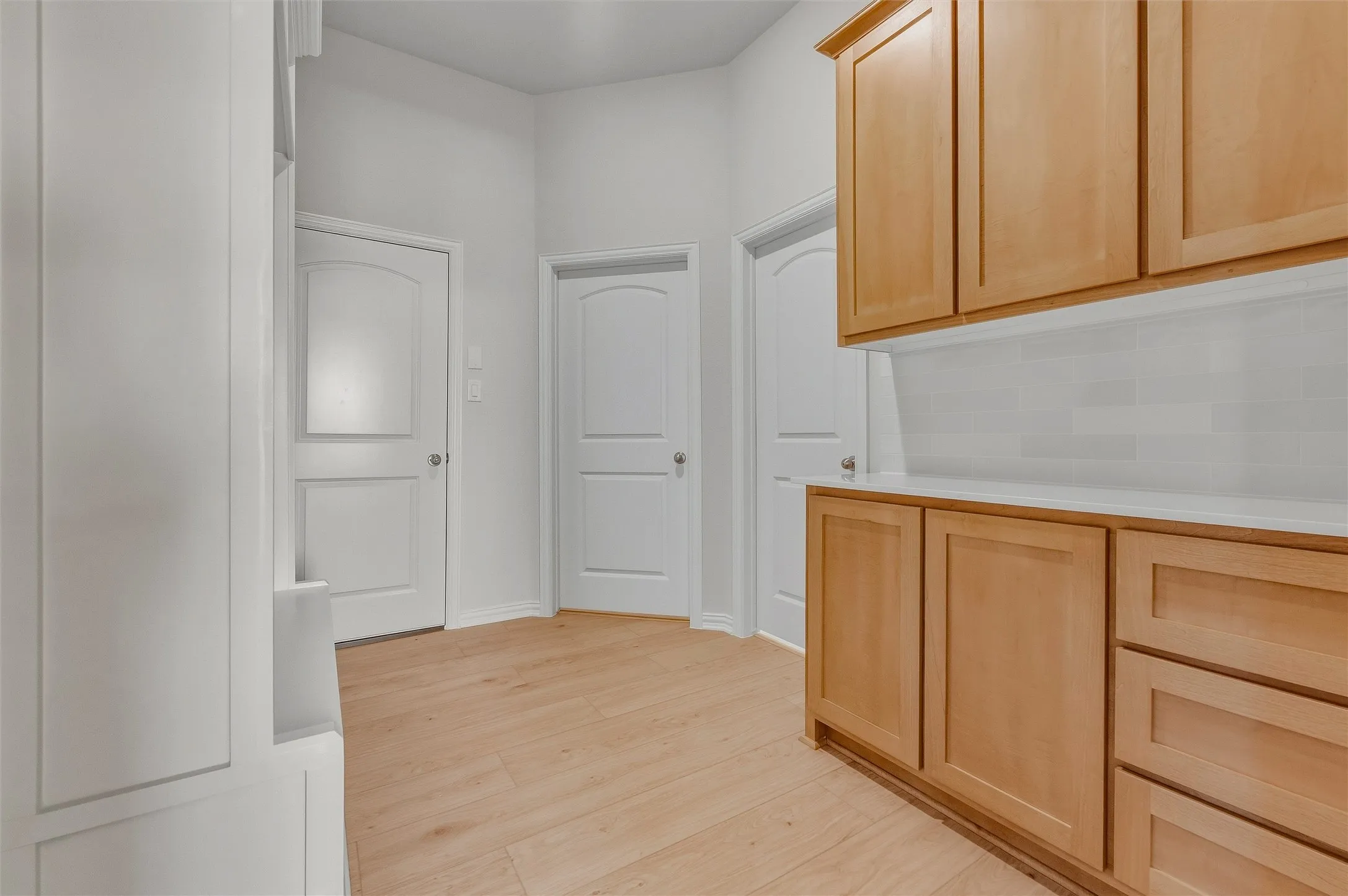 Light wood-finish flooring, built-in cabinetry with light wood doors, white subway tile backsplash, white countertops, and white paneled doors