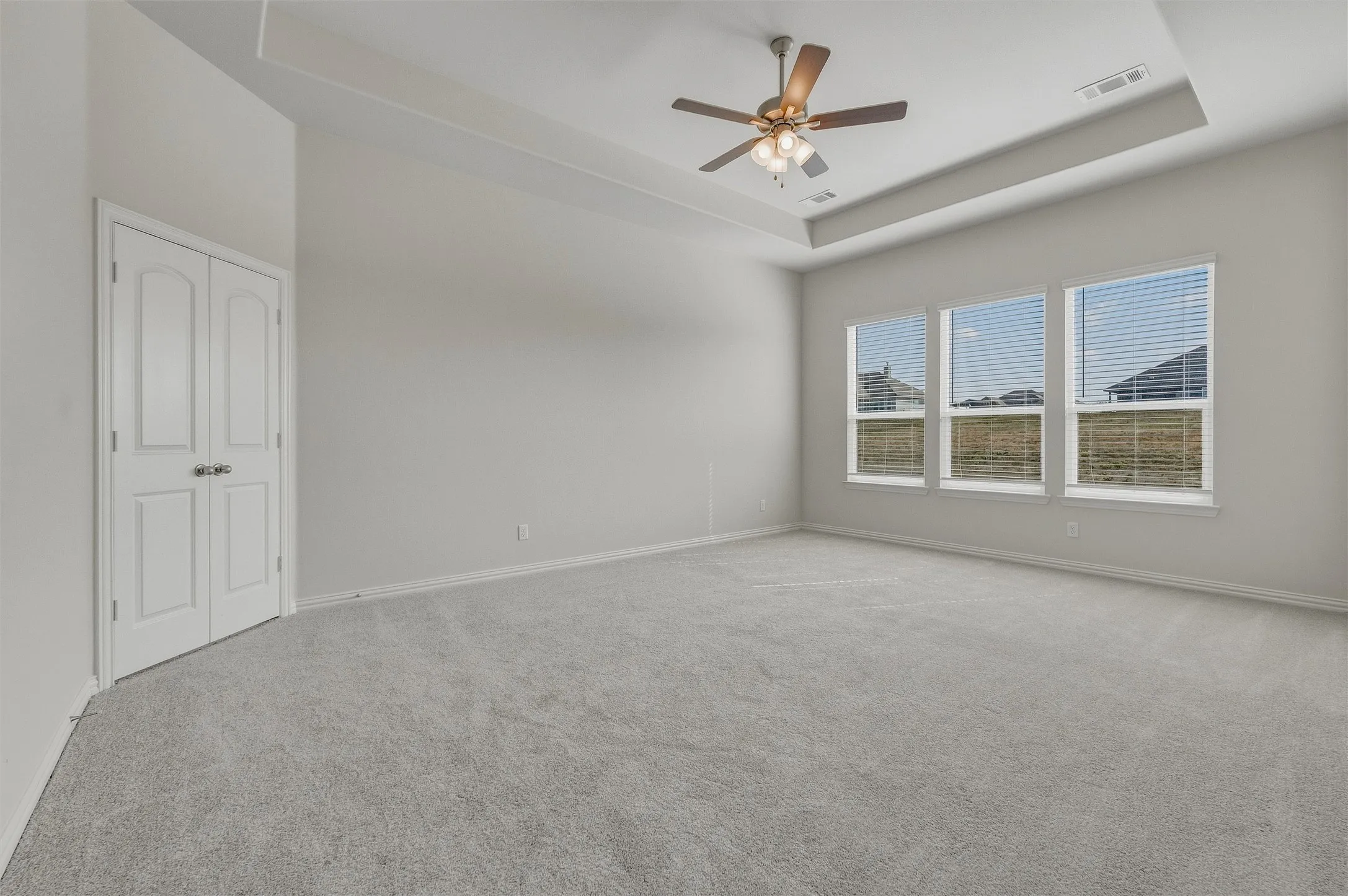 Tray ceiling with a ceiling fan, three windows, light gray carpeting, and white bi-fold doors