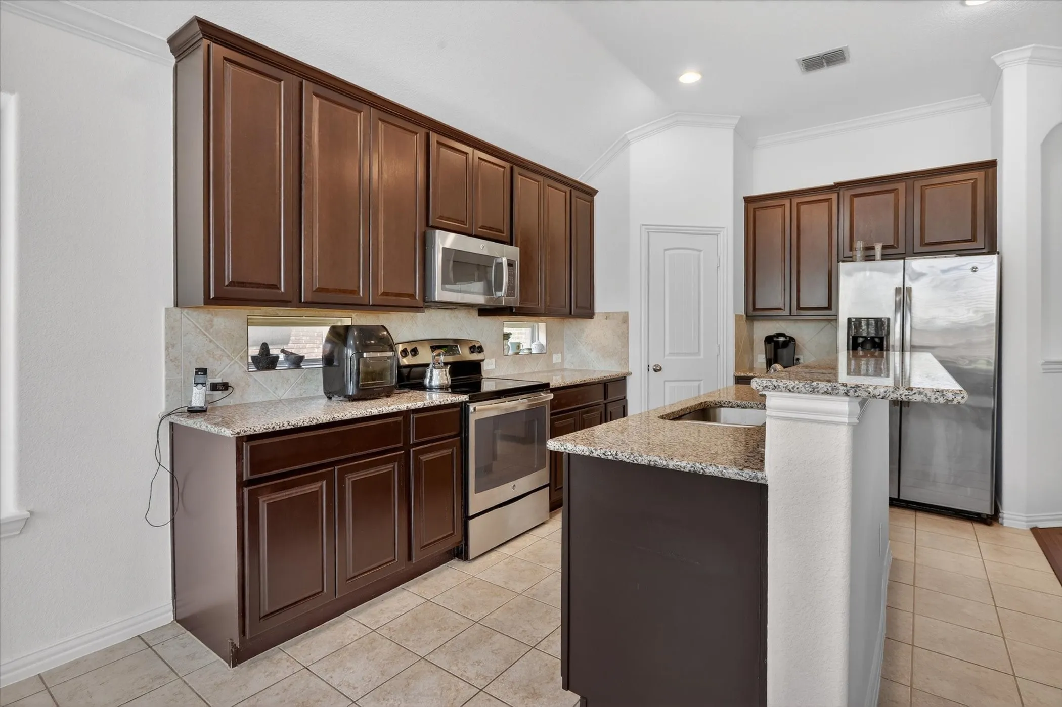 Well-appointed kitchen featuring dark wood cabinetry, granite countertops, stainless steel appliances, a tile backsplash, and tile flooring