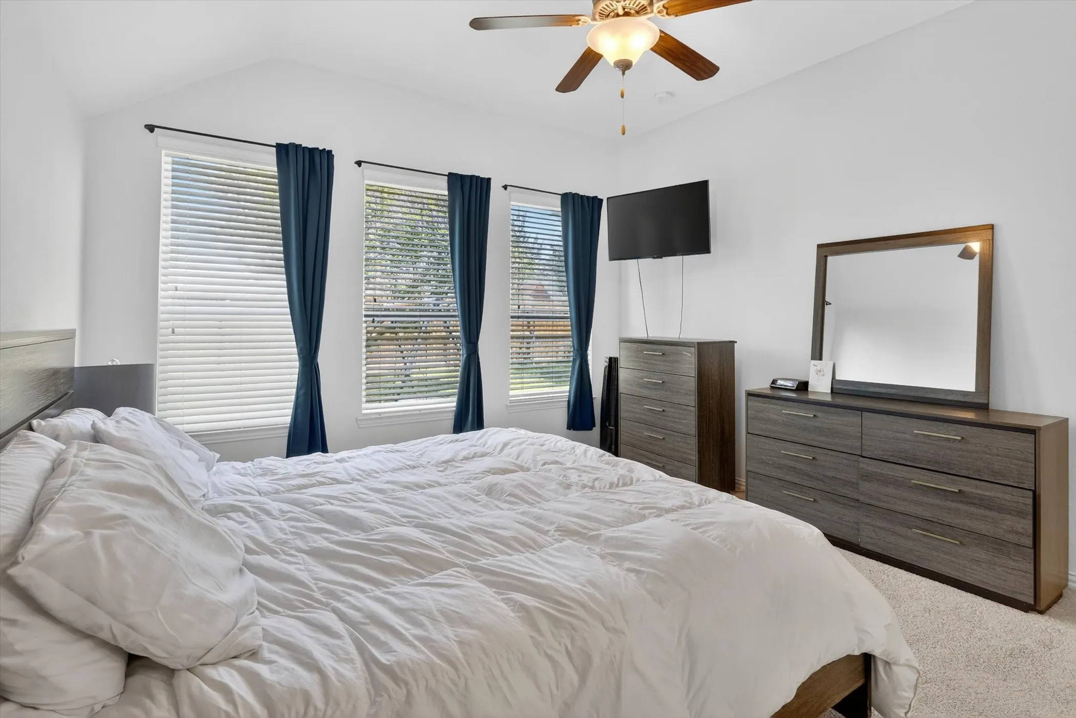 Vaulted ceiling room featuring three windows with blinds, a ceiling fan with light fixture, and neutral-toned carpeting