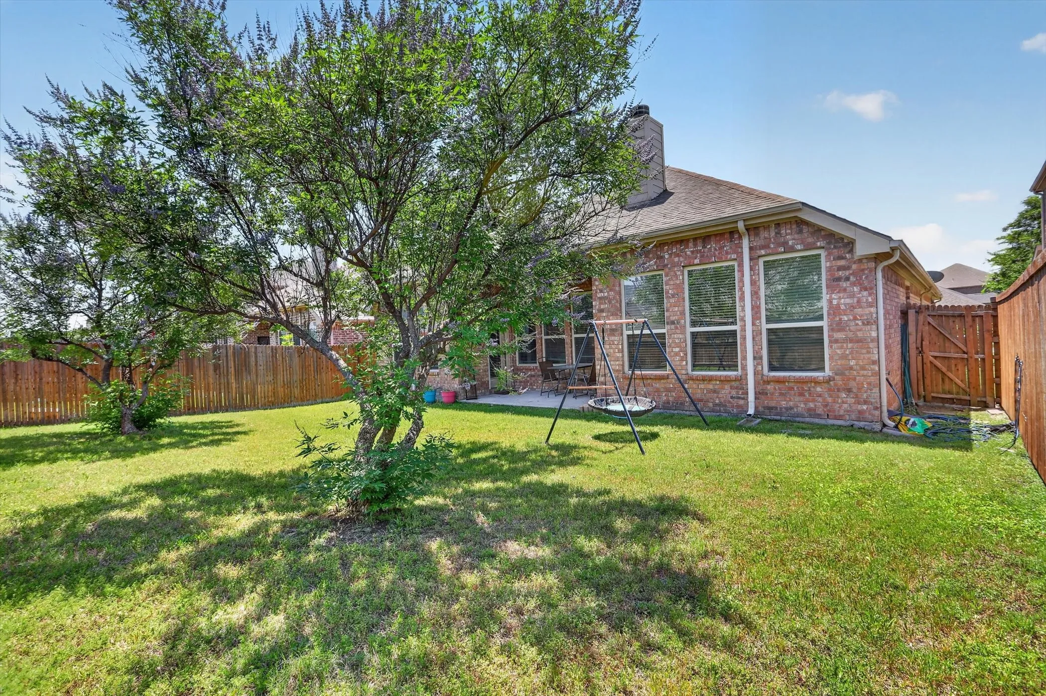 Expansive grassy backyard featuring a mature tree, brick-facade exterior, multiple windows, a wooden privacy fence, and a gate